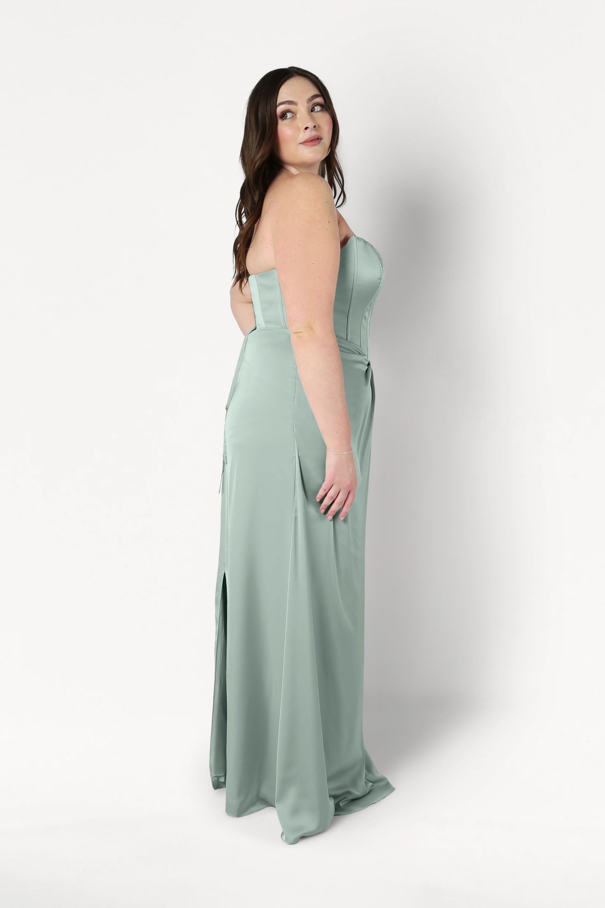 A woman with long brown hair stands sideways against a white background, wearing the size-inclusive TH&TH Selena Bridesmaid Dress in Dusty Eucalyptus Satin with a strapless corset bodice and front slit.