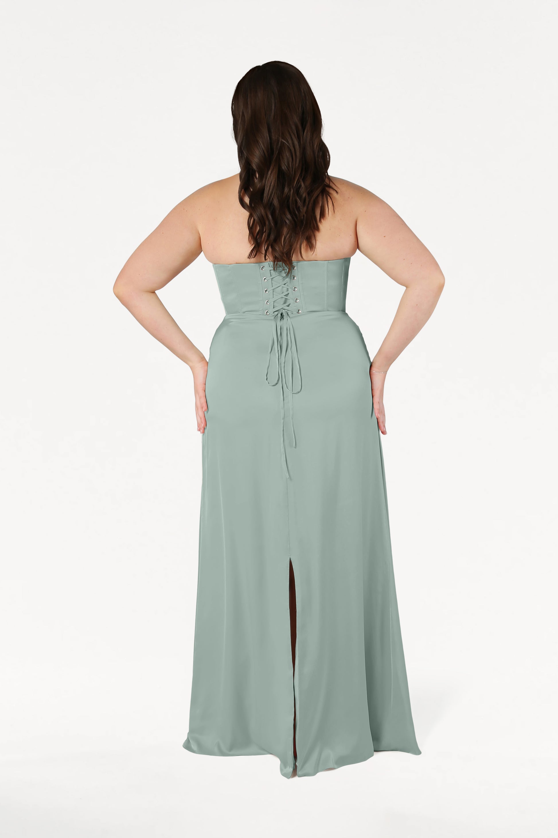 A woman with long brown hair stands facing away, wearing the TH&TH Selena Bridesmaid Dress in Dusty Eucalyptus Satin— a size-inclusive gown with a lace-up corset bodice and bottom slit. The background is plain white.