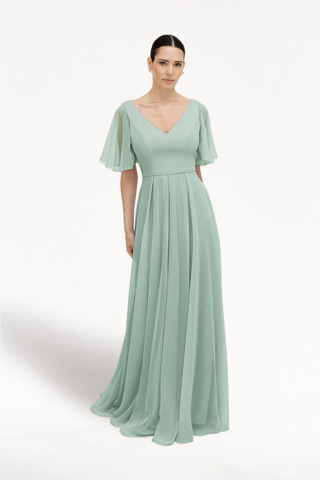 A woman in the TH&TH Phoebe Bridesmaid Dress in Dusty Eucalyptus chiffon, featuring flutter sleeves, a V-neckline, and a pleated floor-length skirt, stands against a plain white background.