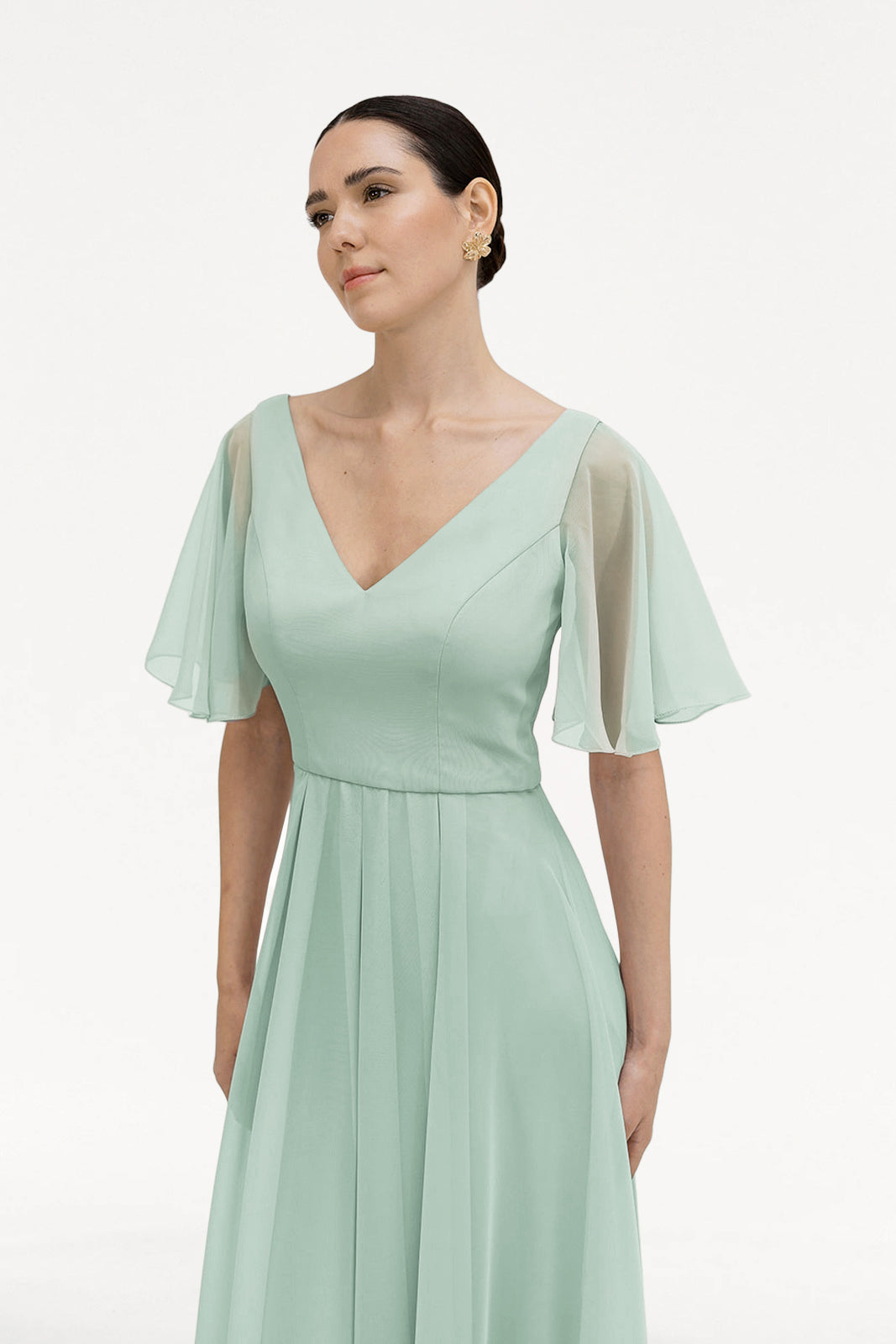 A woman wearing the TH&TH Phoebe Bridesmaid Dress in Dusty Eucalyptus chiffon, featuring sheer flutter sleeves and a V-neckline, stands against a plain white background, looking slightly to the side.