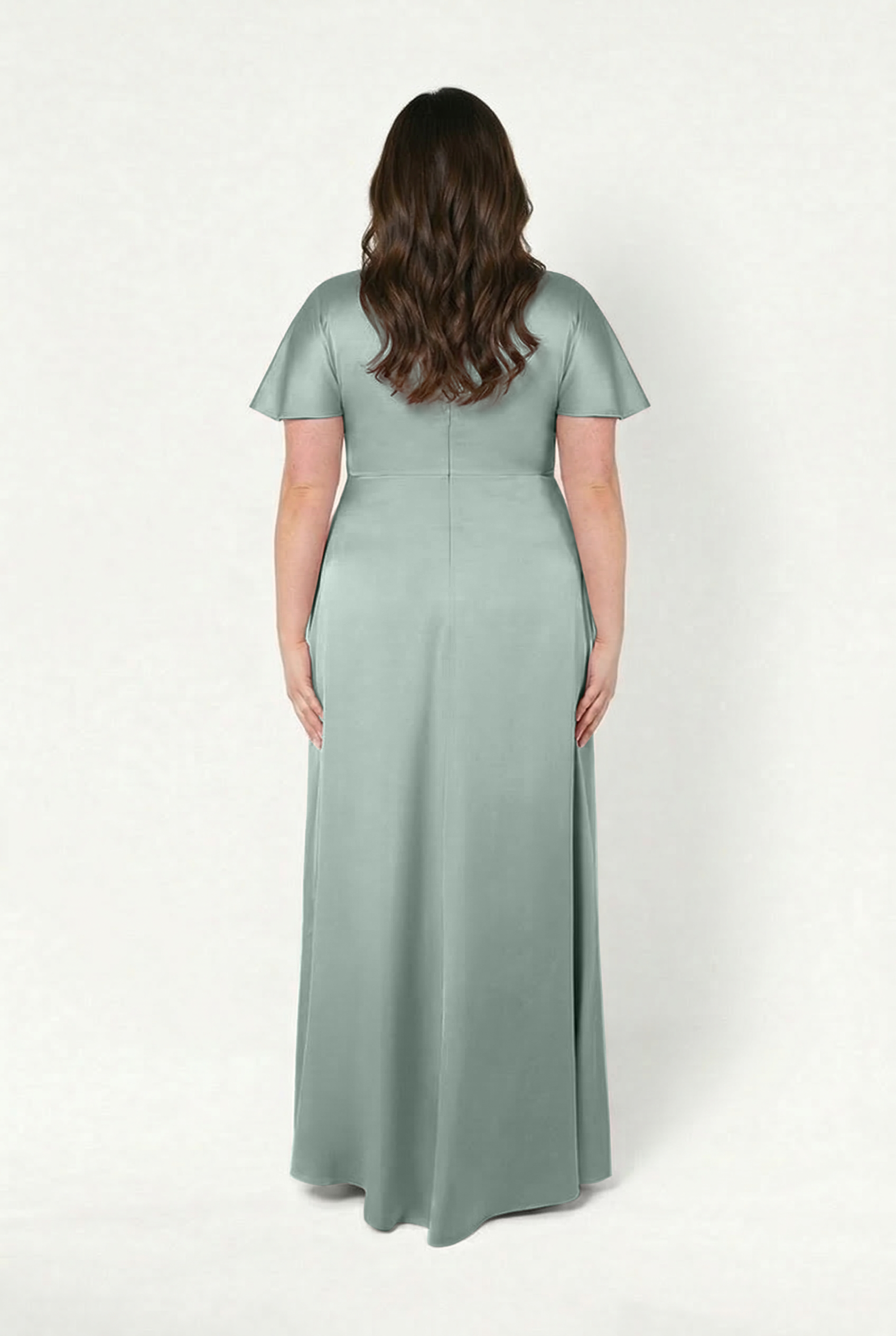 A woman with long, wavy brown hair stands facing away, wearing the TH&TH Camilla Bridesmaid Dress in floor-length, short-sleeved Dusty Eucalyptus satin with a twist-knot bodice against a white background.