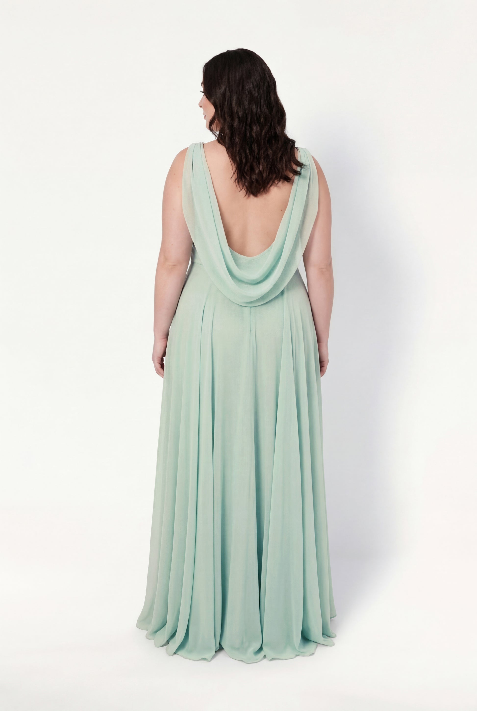 A woman with wavy dark hair stands with her back to the camera, wearing the TH&TH Athena Bridesmaid Dress in Dusty Eucalyptus—a flowing chiffon gown with a low cowl draped back—set against a plain white background.