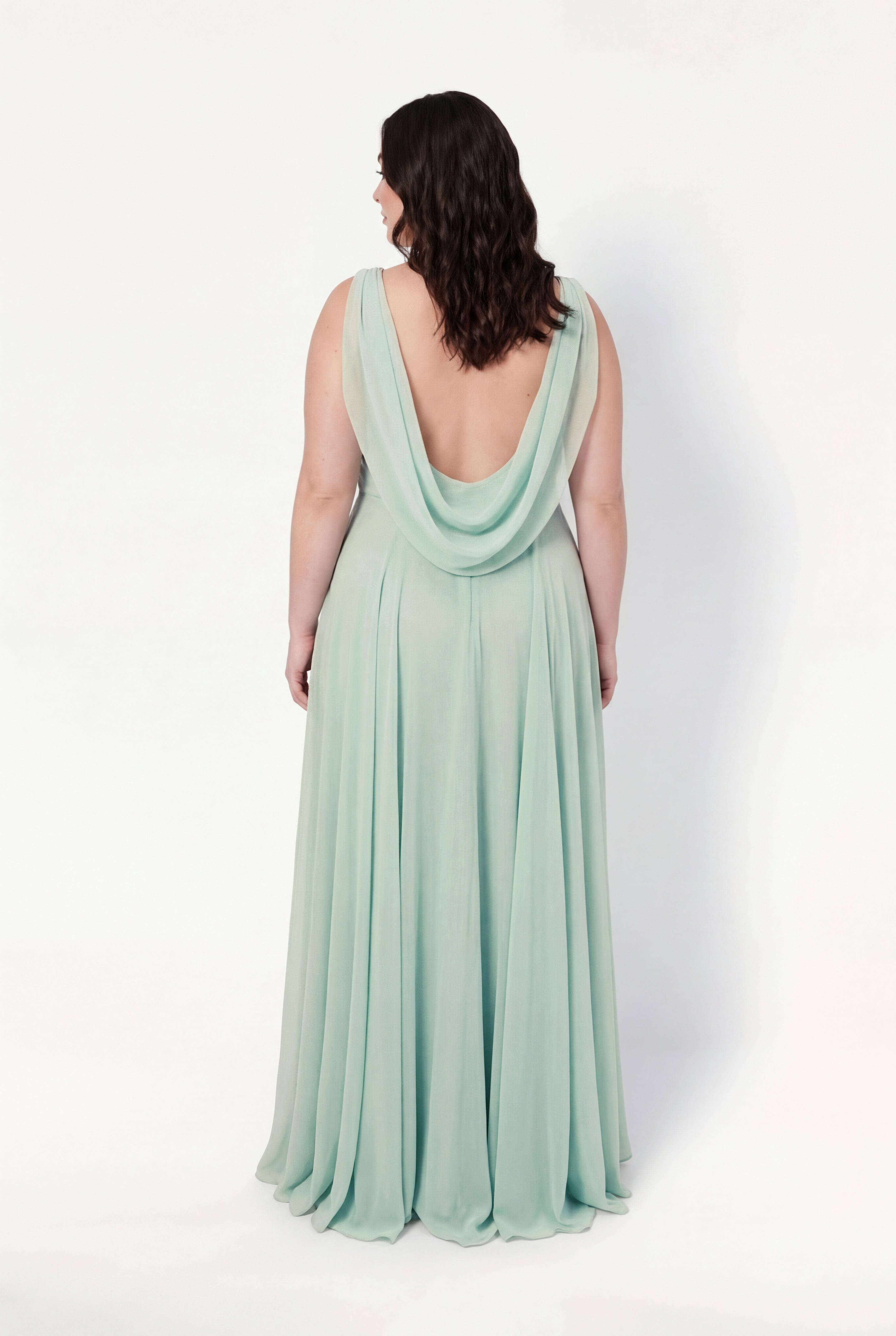 A woman with wavy dark hair stands with her back to the camera, wearing the TH&TH Athena Bridesmaid Dress in Dusty Eucalyptus—a flowing chiffon gown with a low cowl draped back—set against a plain white background.