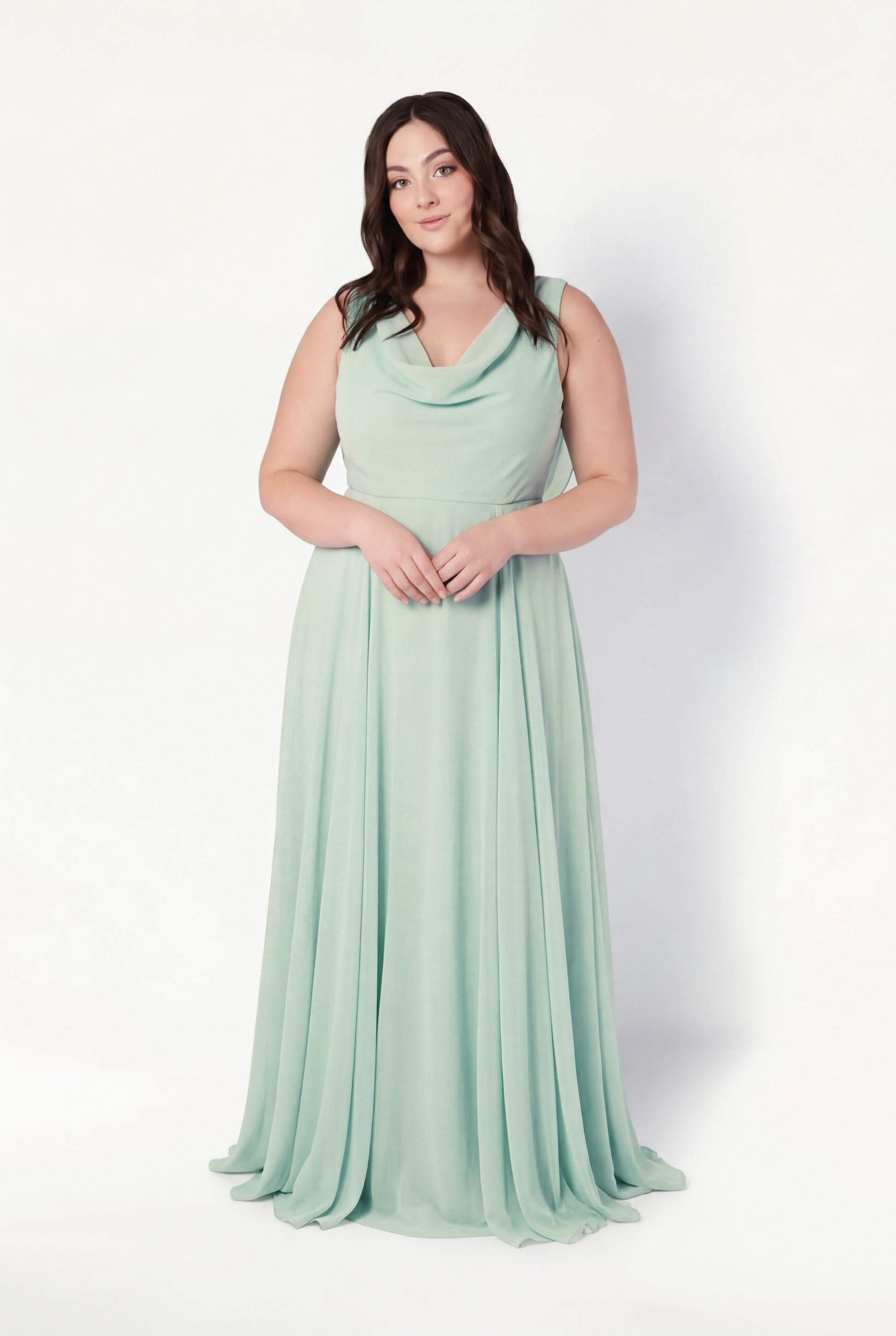 A woman with long, dark hair stands against a white background, wearing the TH&TH Athena Bridesmaid Dress in Dusty Eucalyptus—a sleeveless, floor-length chiffon gown with a cowl neck and flowing skirt, perfect for bridesmaids.