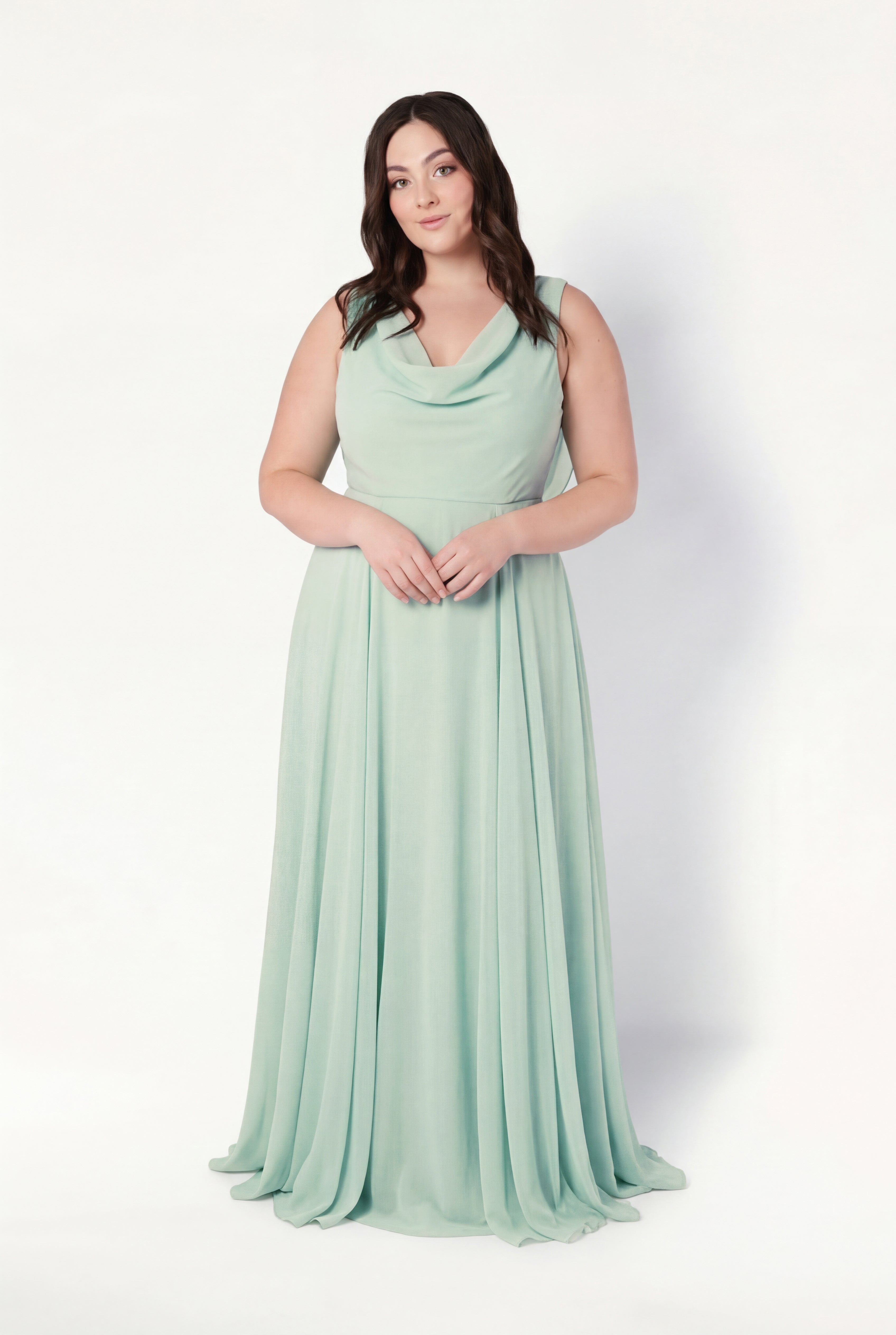 A woman with long, dark hair stands against a white background, wearing the TH&TH Athena Bridesmaid Dress in Dusty Eucalyptus—a sleeveless, floor-length chiffon gown with a cowl neck and flowing skirt, perfect for bridesmaids.