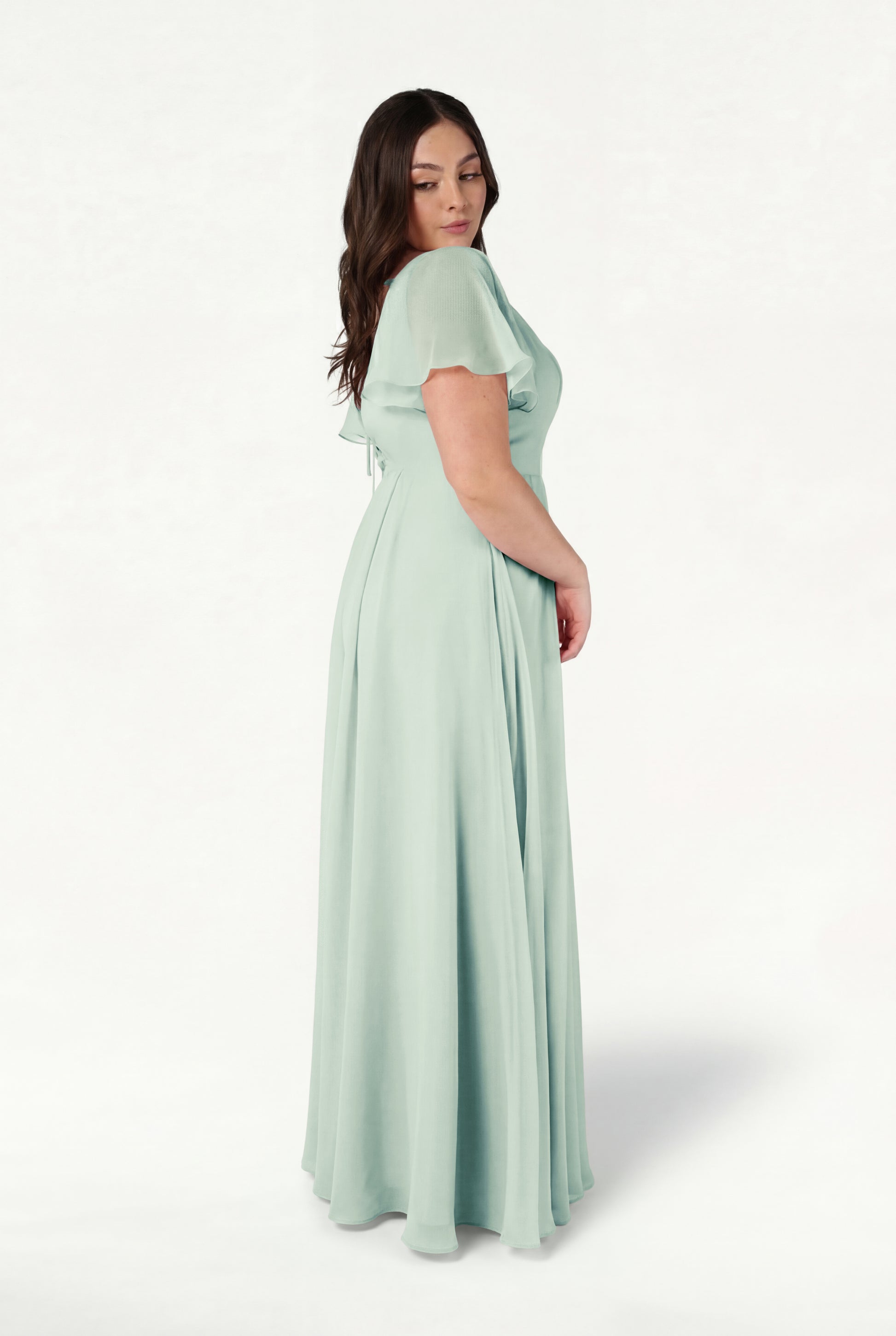 A woman with long dark hair stands sideways, wearing the TH&TH Phoebe Bridesmaid Dress in Dusty Eucalyptus chiffon—a floor-length, bump-friendly gown with a fitted waist and short sleeves—against a plain white background.