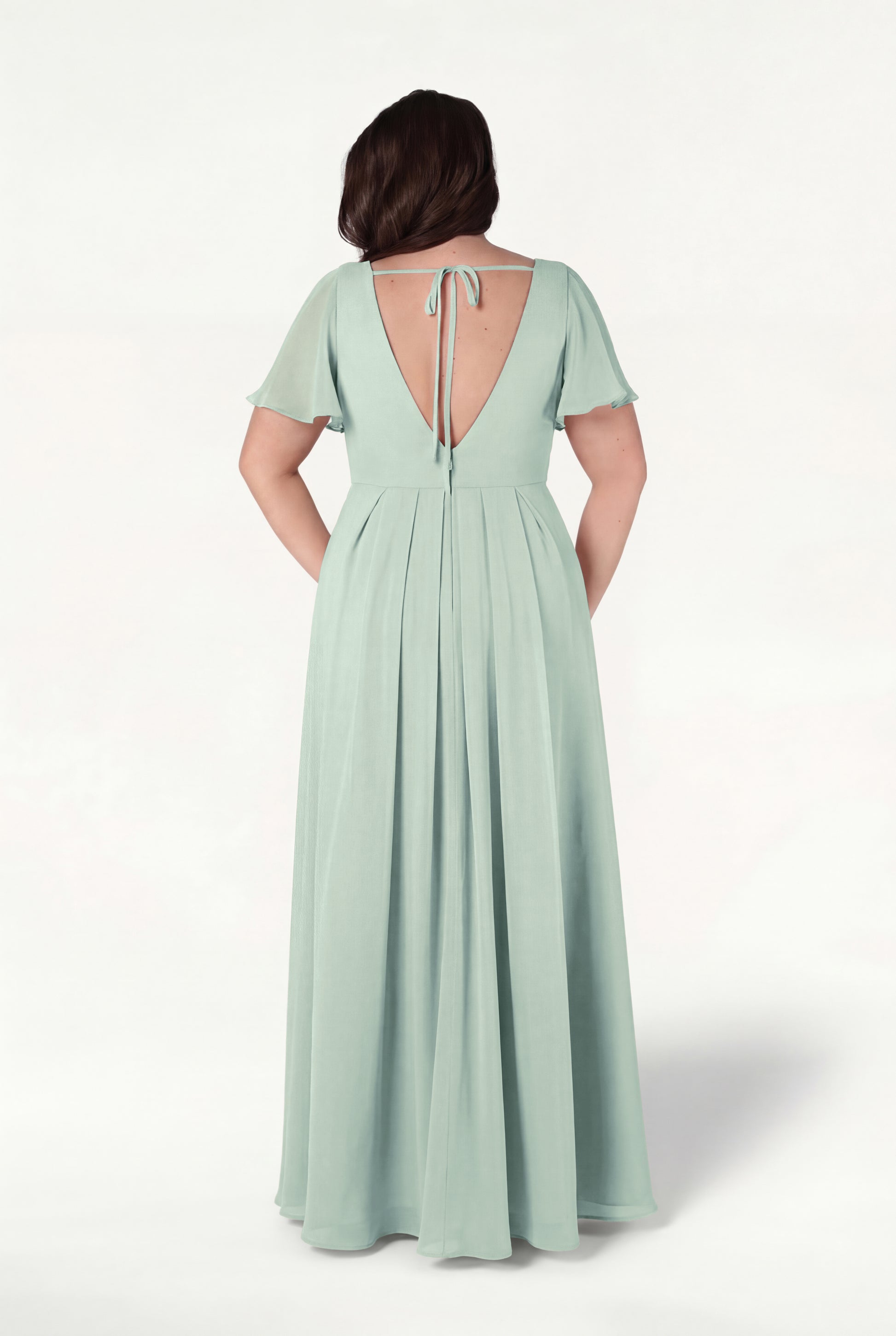 A woman faces away, wearing the TH&TH Phoebe Bridesmaid Dress in Dusty Eucalyptus chiffon—long, flowing with short flutter sleeves, a deep V-back, and tie detail. The background is plain white.