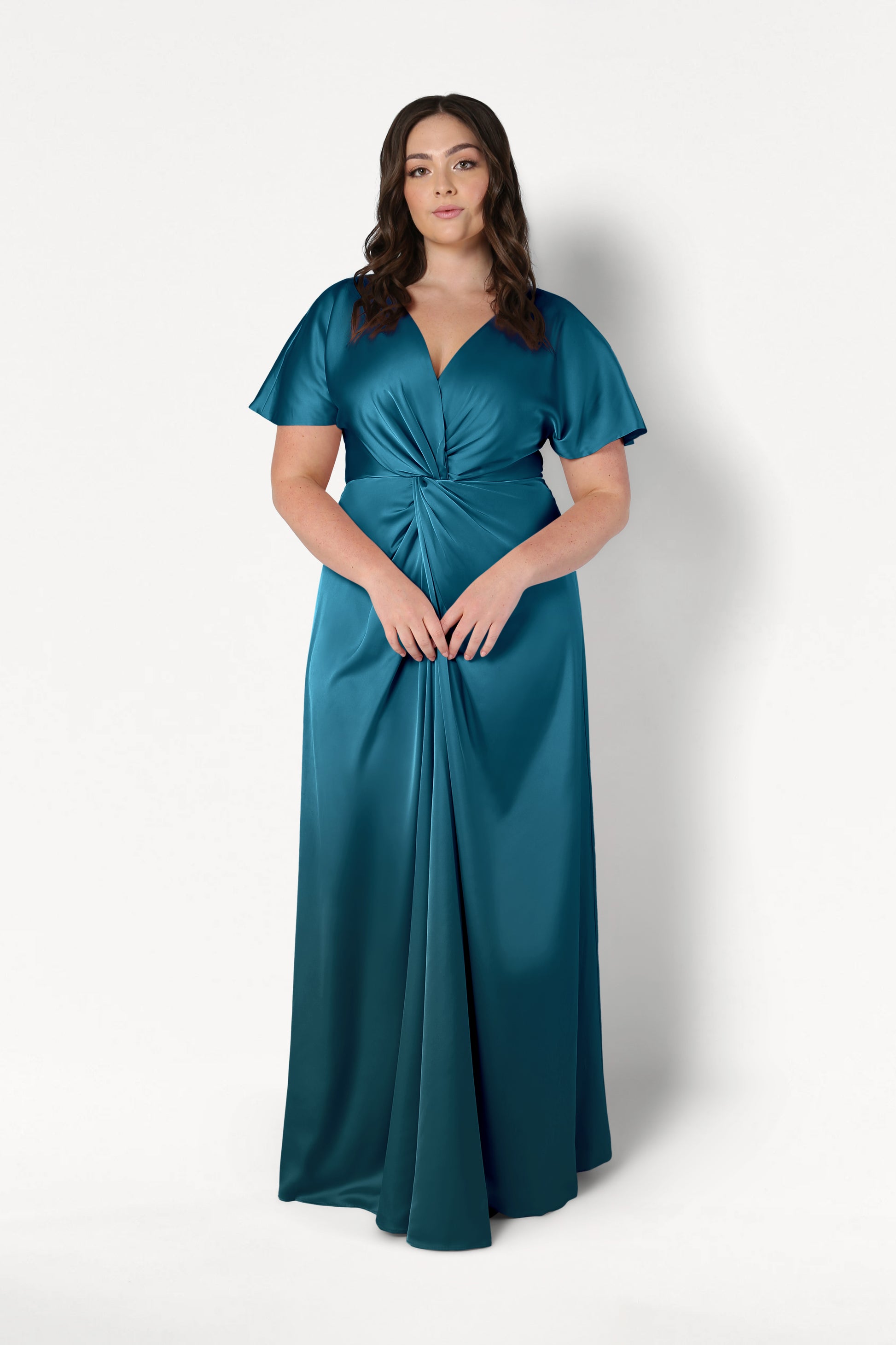 A woman with long brown hair wears the Camilla Bridesmaid Dress in Emerald Dawn Satin by TH&TH— a modern, floor-length gown with short flutter sleeves and a gathered twist waist—standing against a plain white background.