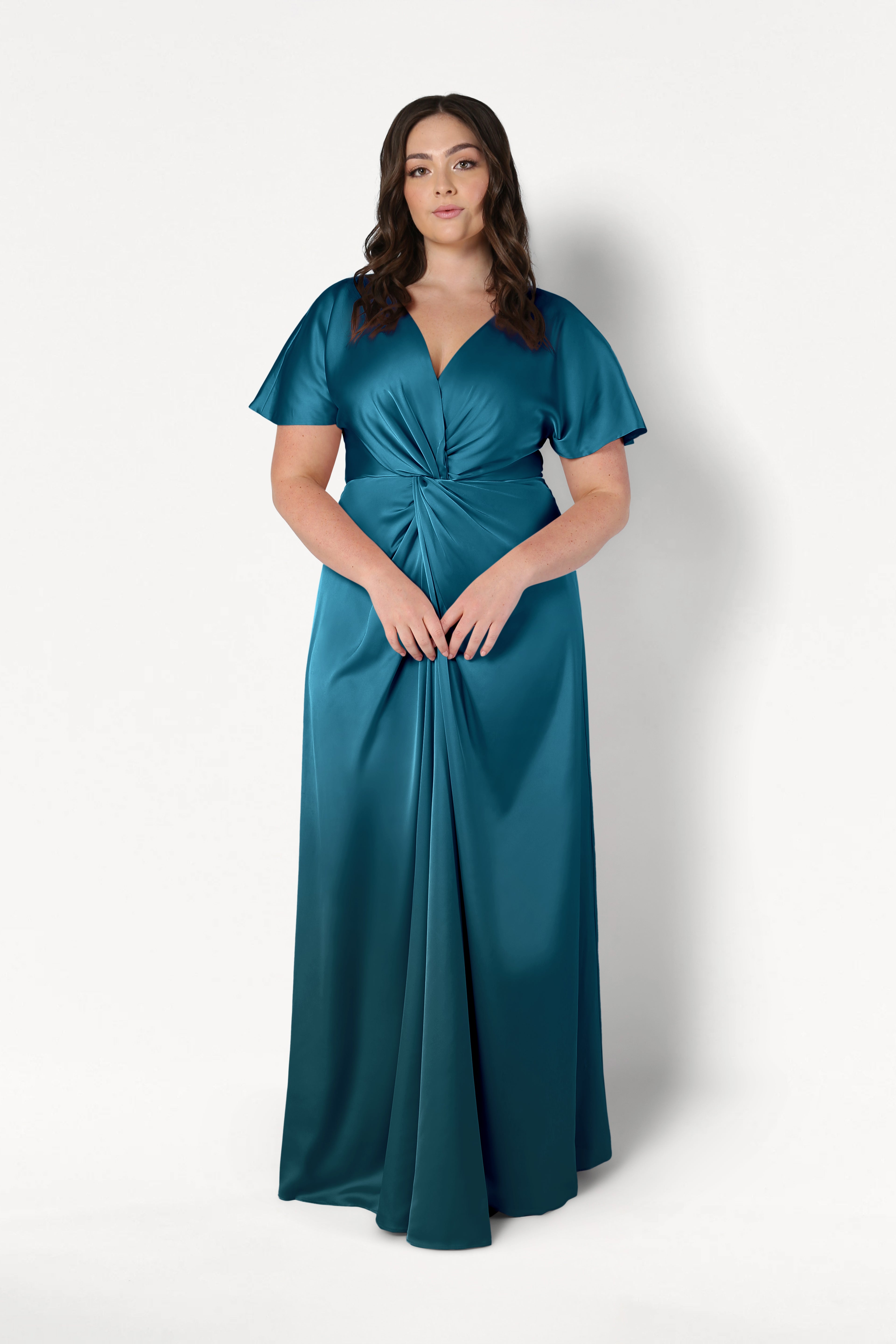 A woman with long brown hair wears the Camilla Bridesmaid Dress in Emerald Dawn Satin by TH&TH— a modern, floor-length gown with short flutter sleeves and a gathered twist waist—standing against a plain white background.