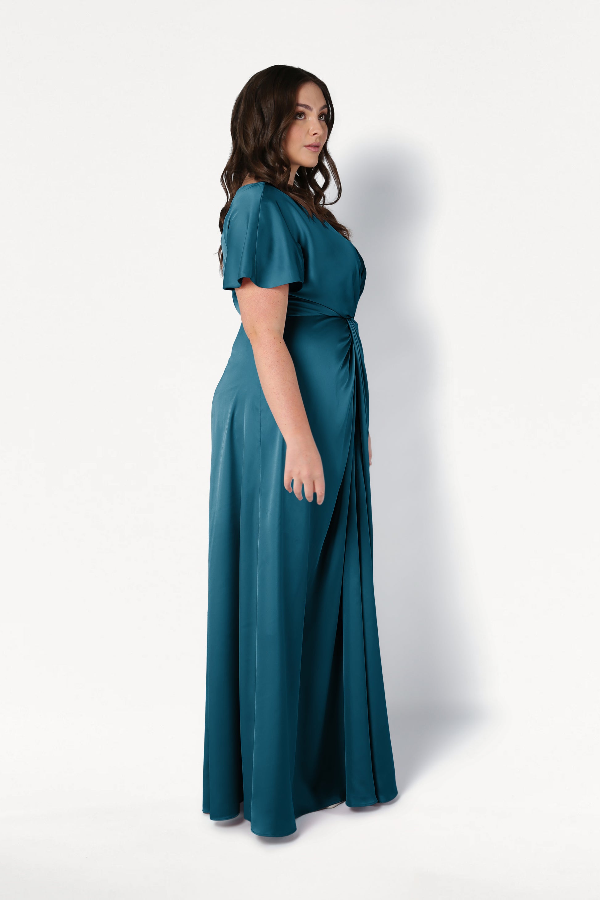 A woman with wavy brown hair stands sideways in the TH&TH Camilla Bridesmaid Dress in Emerald Dawn Satin, a floor-length, short-sleeve gown with a gathered waist, set against a plain white background.