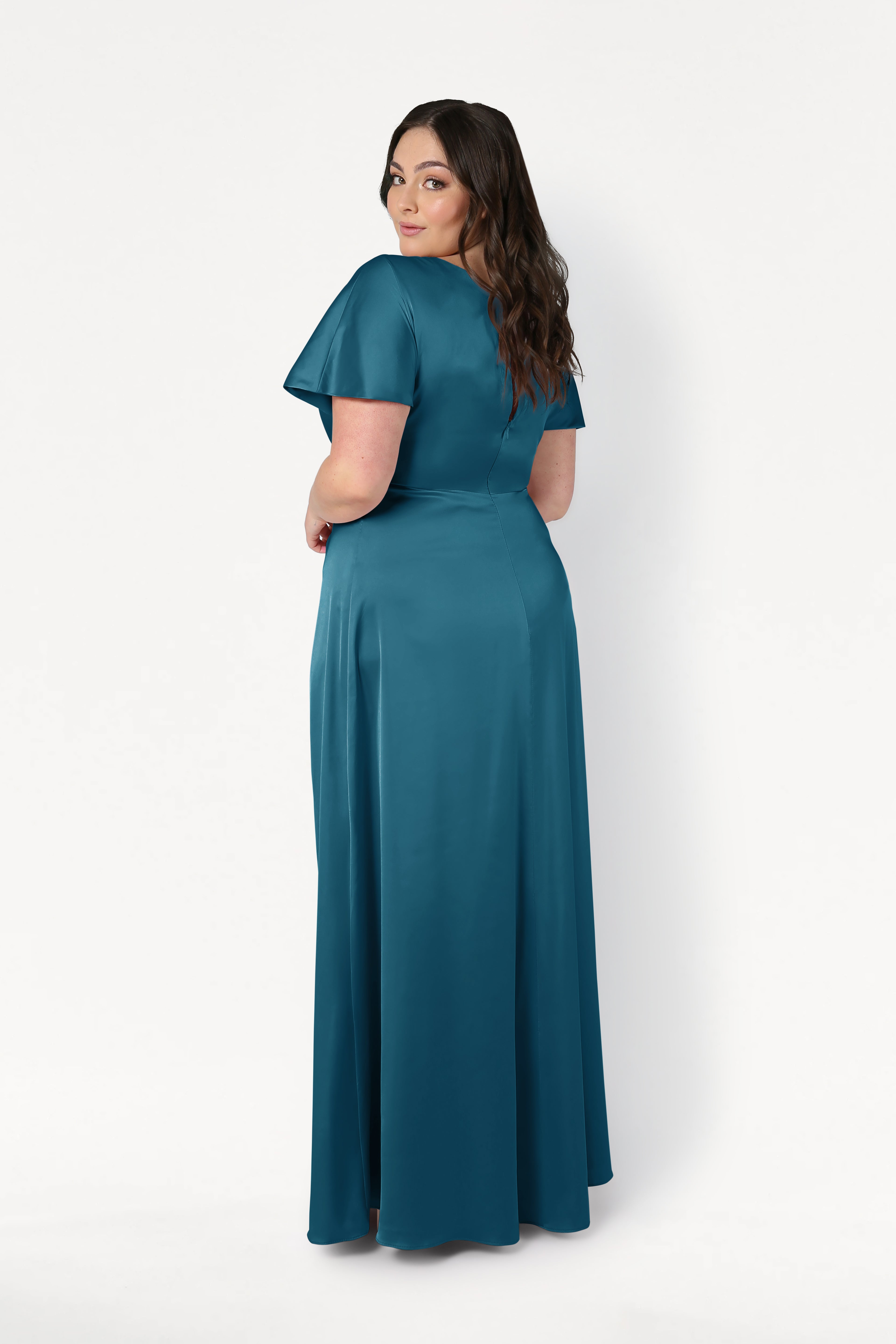 A woman with long dark hair stands, looking over her shoulder in the floor-length Camilla Bridesmaid Dress in Emerald Dawn Satin by TH&TH. The dress has short sleeves and is set against a plain white background.