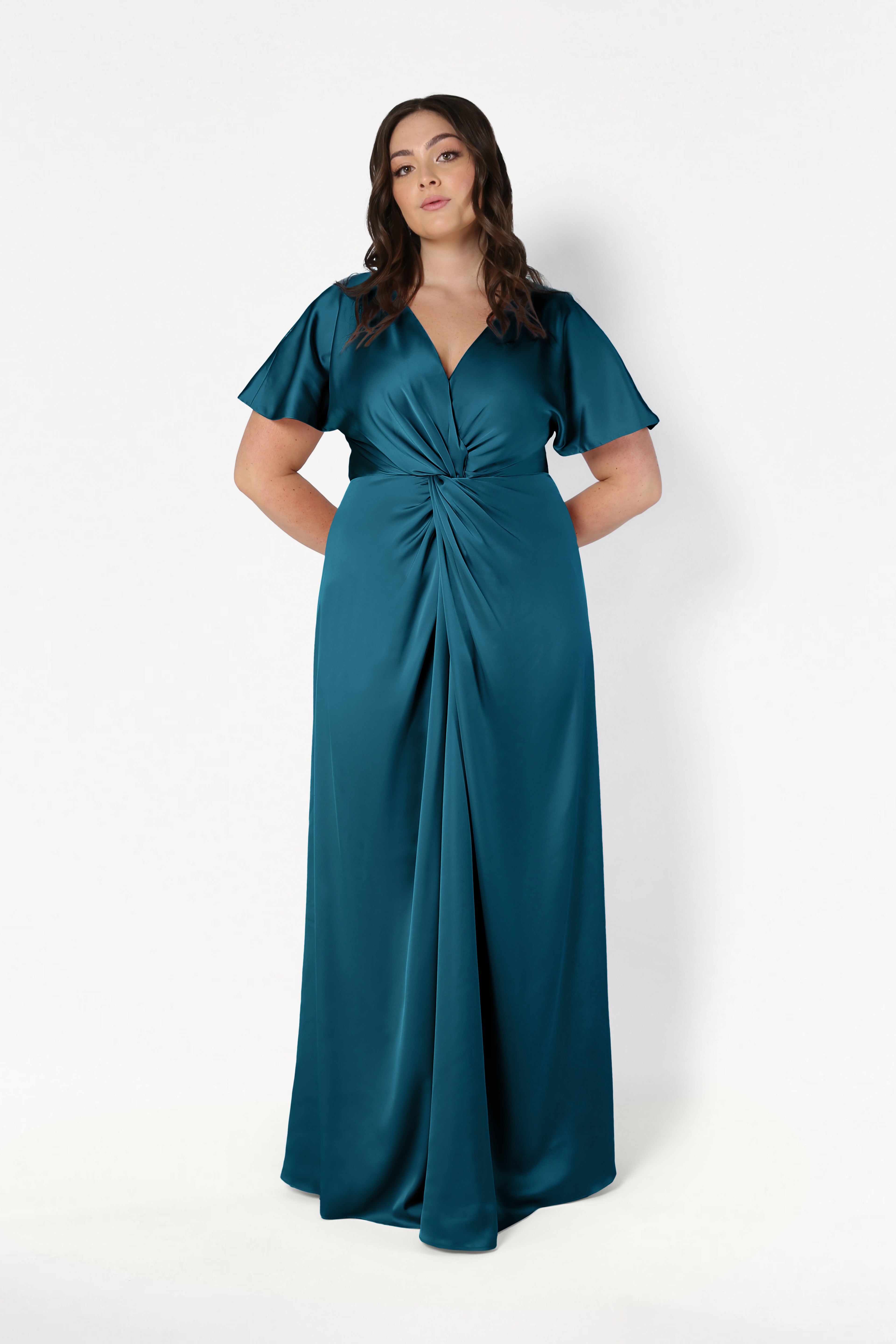 A woman with long wavy hair wears the TH&TH Camilla Bridesmaid Dress in Emerald Dawn Satin, featuring short flutter sleeves, a V-neckline, and a gathered twist waist, standing against a plain white background.