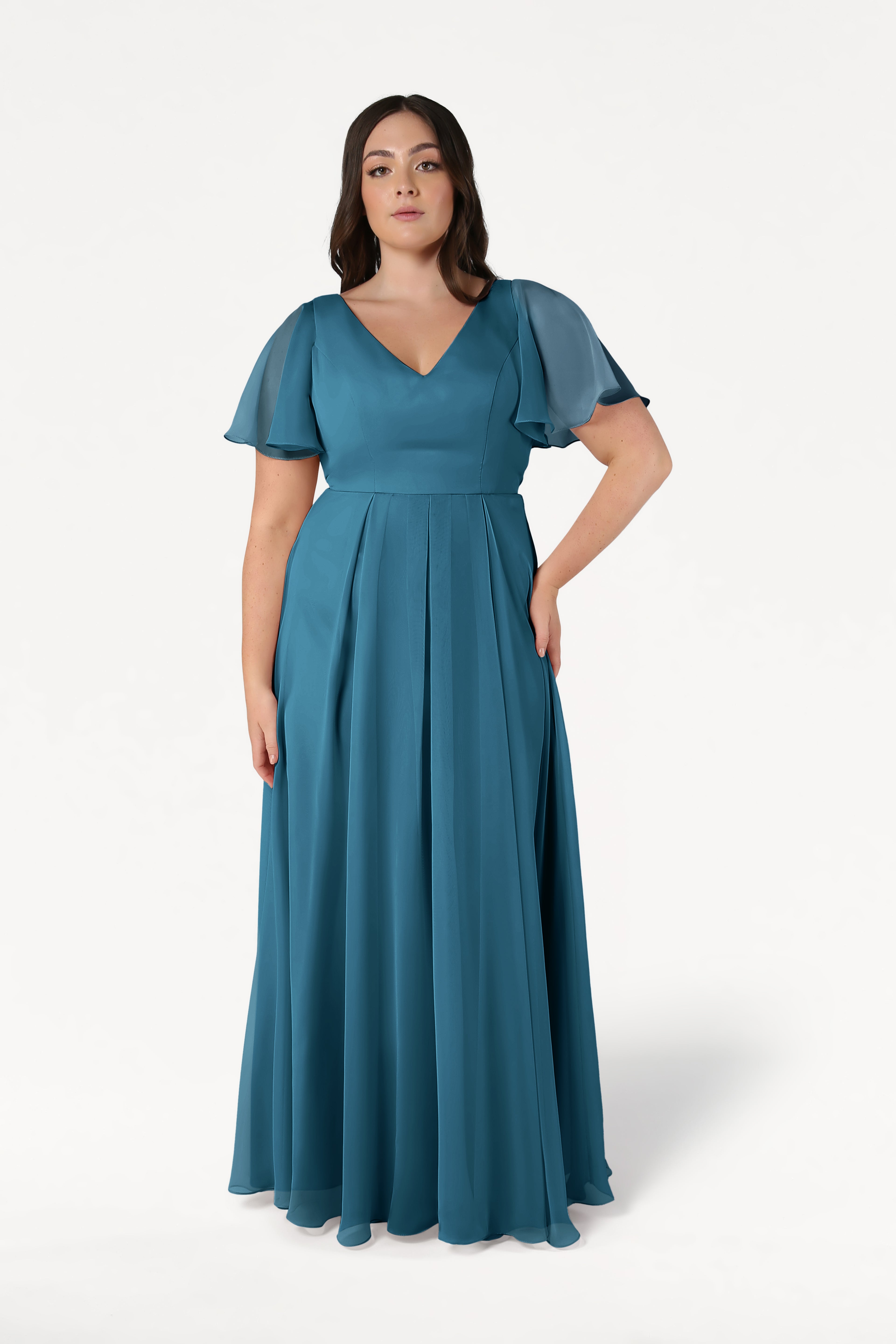 A woman poses against a white background in the TH&TH Phoebe Bridesmaid Dress in Emerald Dawn Chiffon, featuring short sheer flutter sleeves, a V-neckline, pleated floor-length skirt, and a bump-friendly silhouette. She has dark hair and a neutral expression.