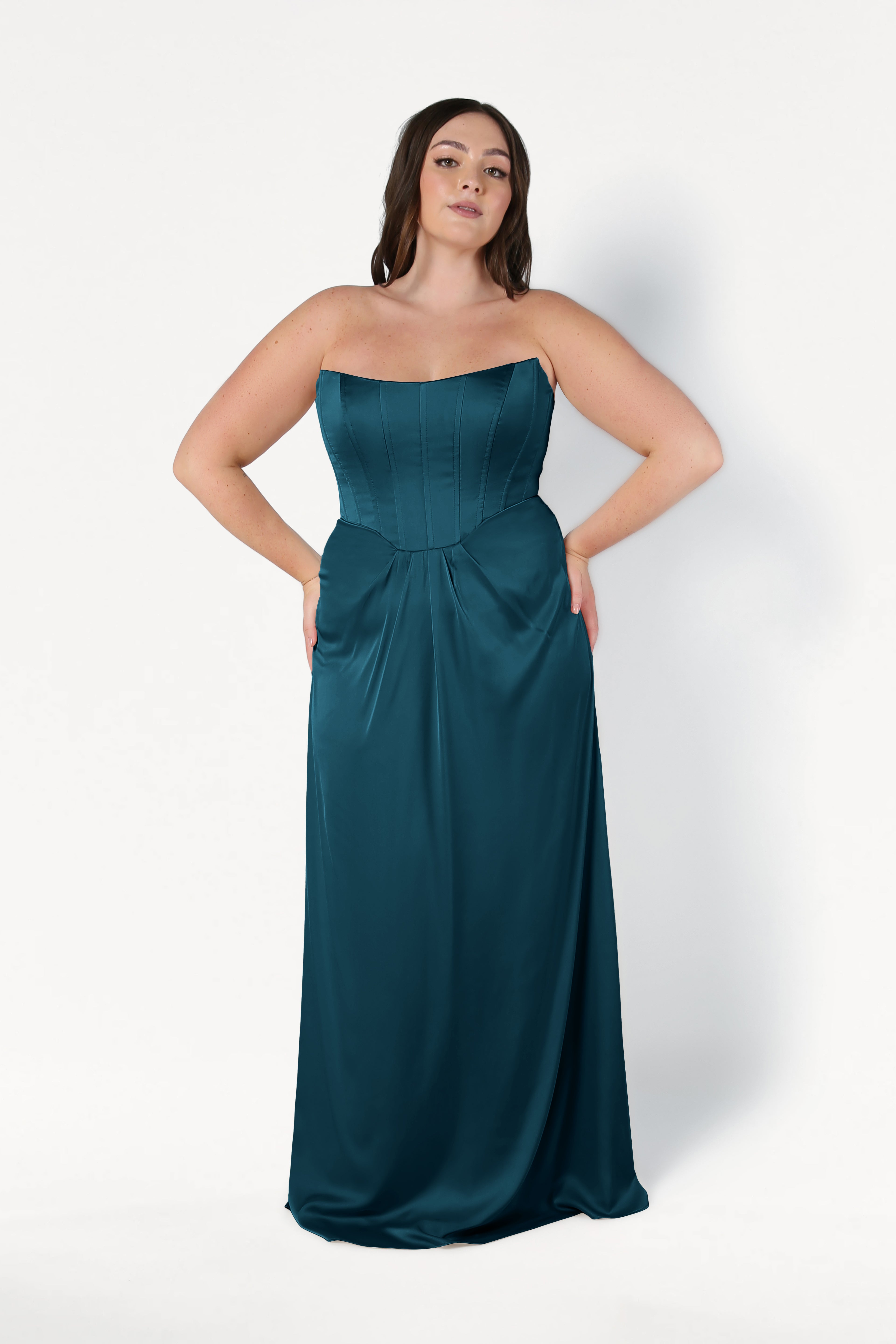 A woman with long dark hair models the TH&TH Selena Bridesmaid Dress in Emerald Dawn Satin, featuring a strapless fitted corset bodice and flowing skirt, posing with hands on her hips against a plain white background.