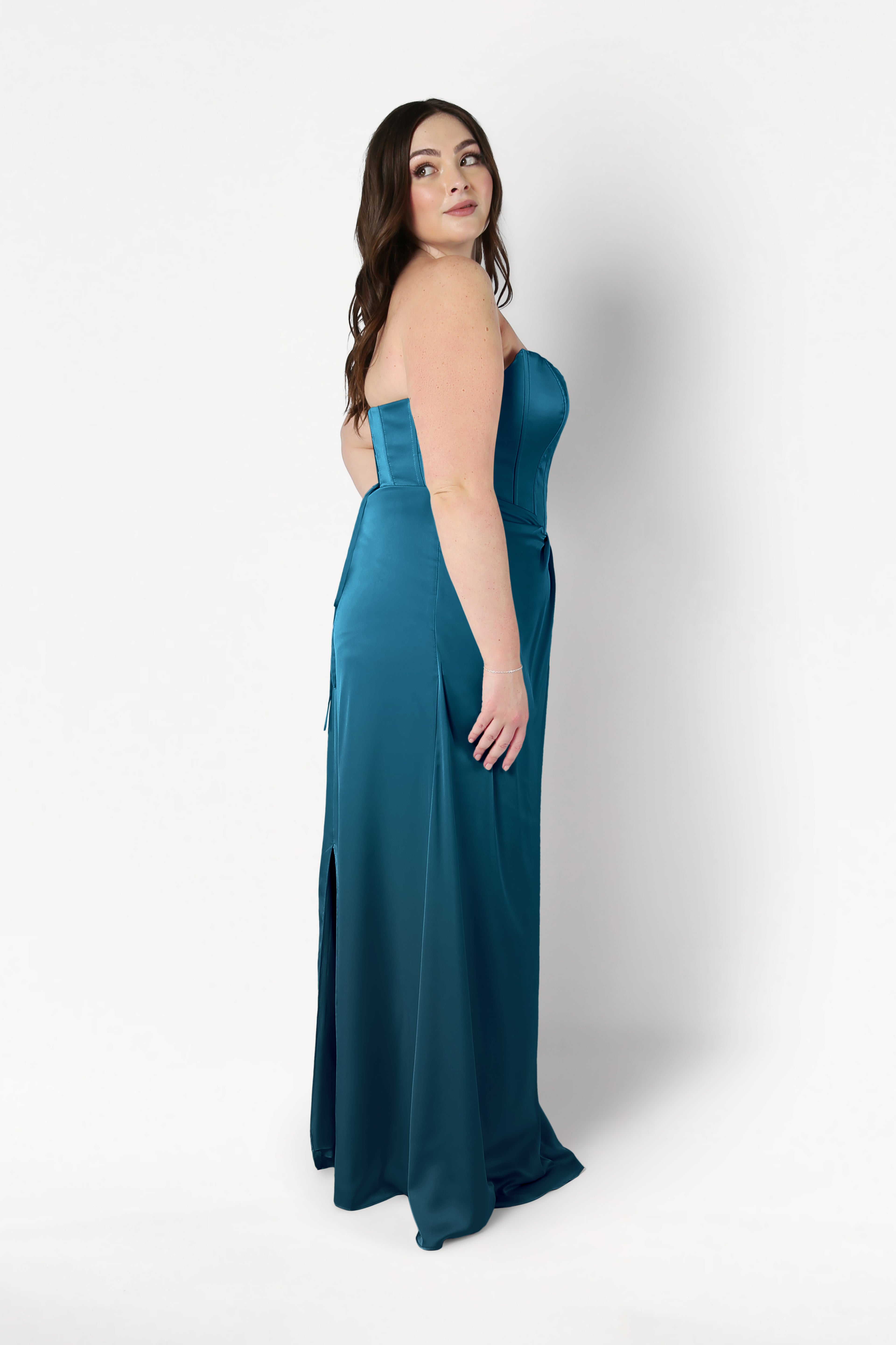 A woman with long brown hair wears the TH&TH Selena Bridesmaid Dress in Emerald Dawn Satin, featuring a strapless corset bodice. She stands sideways, looking over her shoulder against a plain white background.