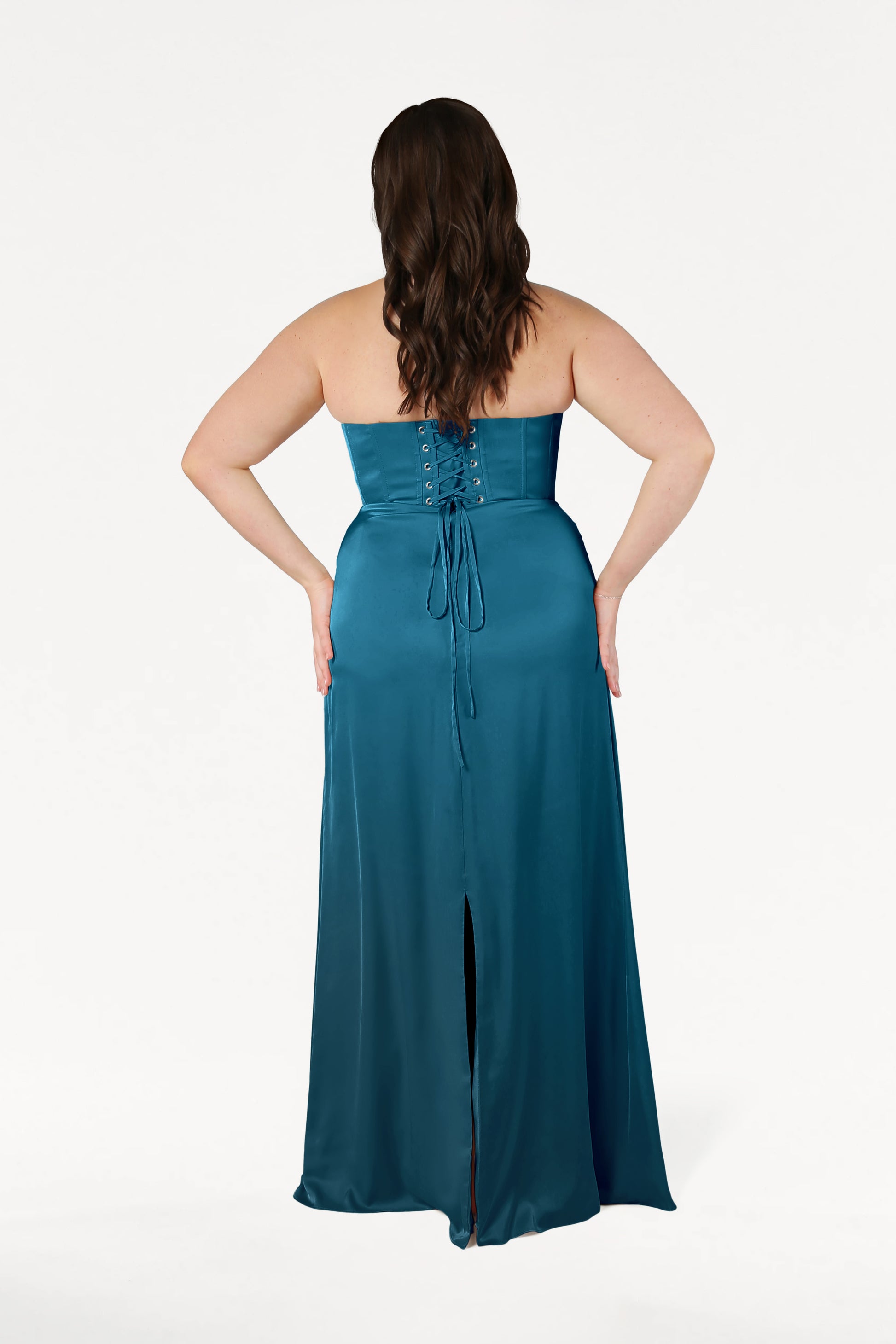 A woman with long brown hair faces away, wearing the size-inclusive TH&TH Selena Bridesmaid Dress in Emerald Dawn Satin, featuring a corset bodice, lace-up back, and back slit. The background is plain white.