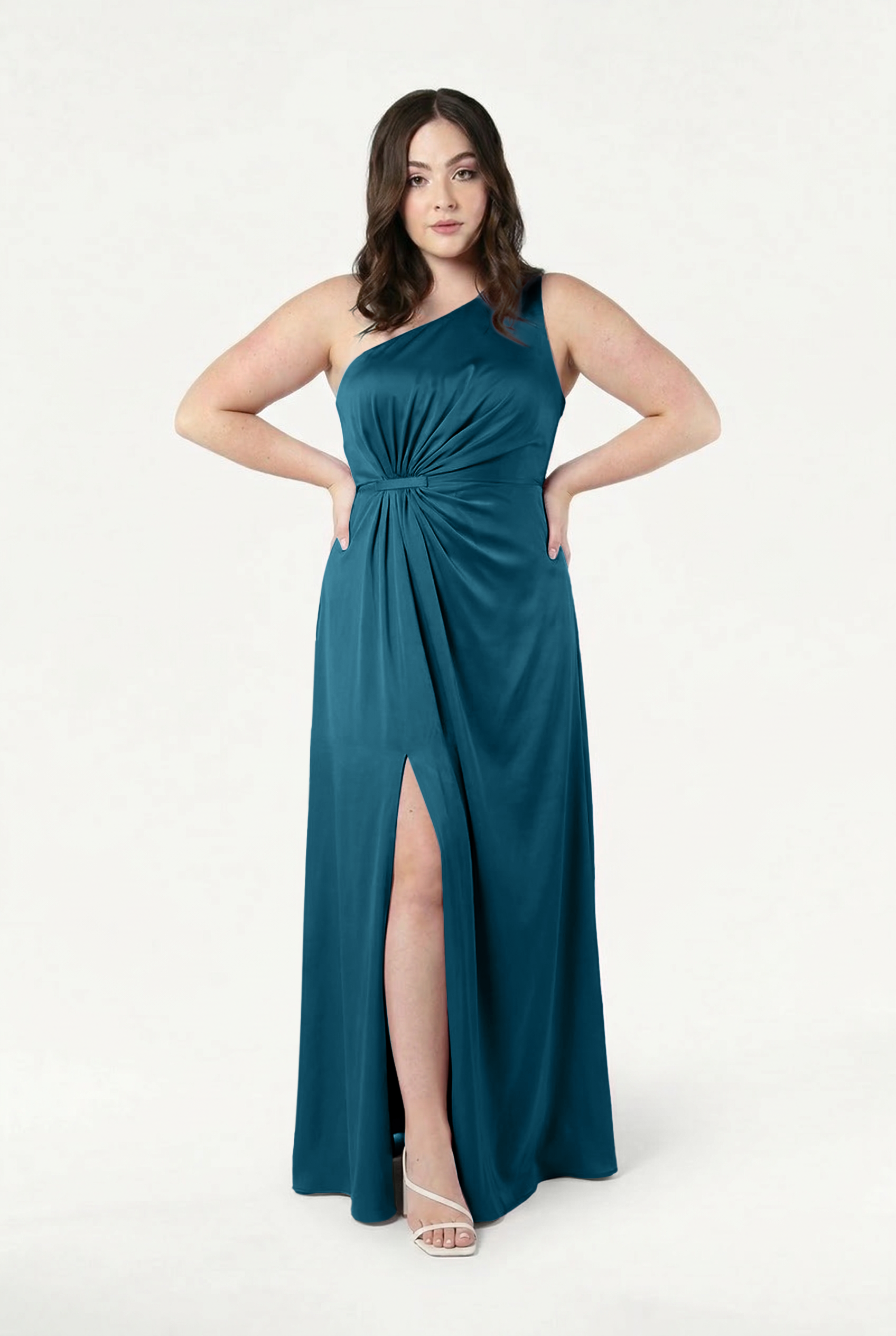 A woman stands confidently against a white background wearing the TH&TH Greta Bridesmaid Dress in Emerald Dawn Satin—a floor-length, teal one-shoulder gown with waist knot detail, thigh-high slit, and paired with white strappy sandals.
