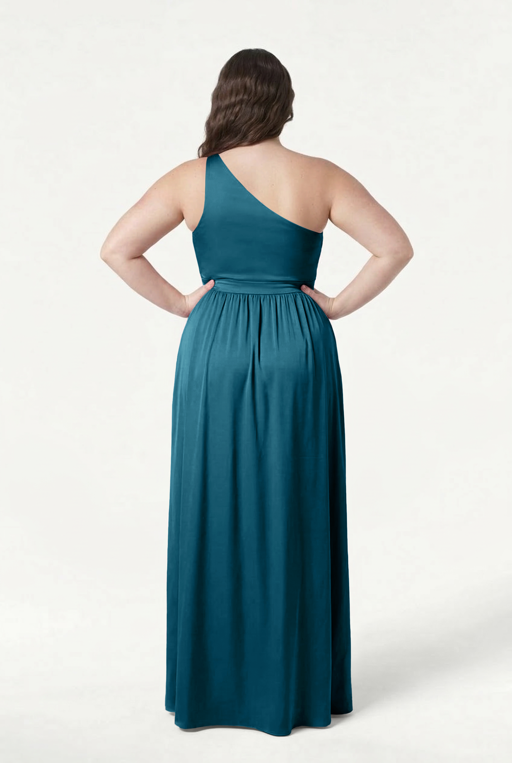 A woman stands hands on hips in the TH&TH Greta Bridesmaid Dress in Emerald Dawn Satin, photographed from behind to showcase its flowing skirt and fitted waistband against a plain white background.
