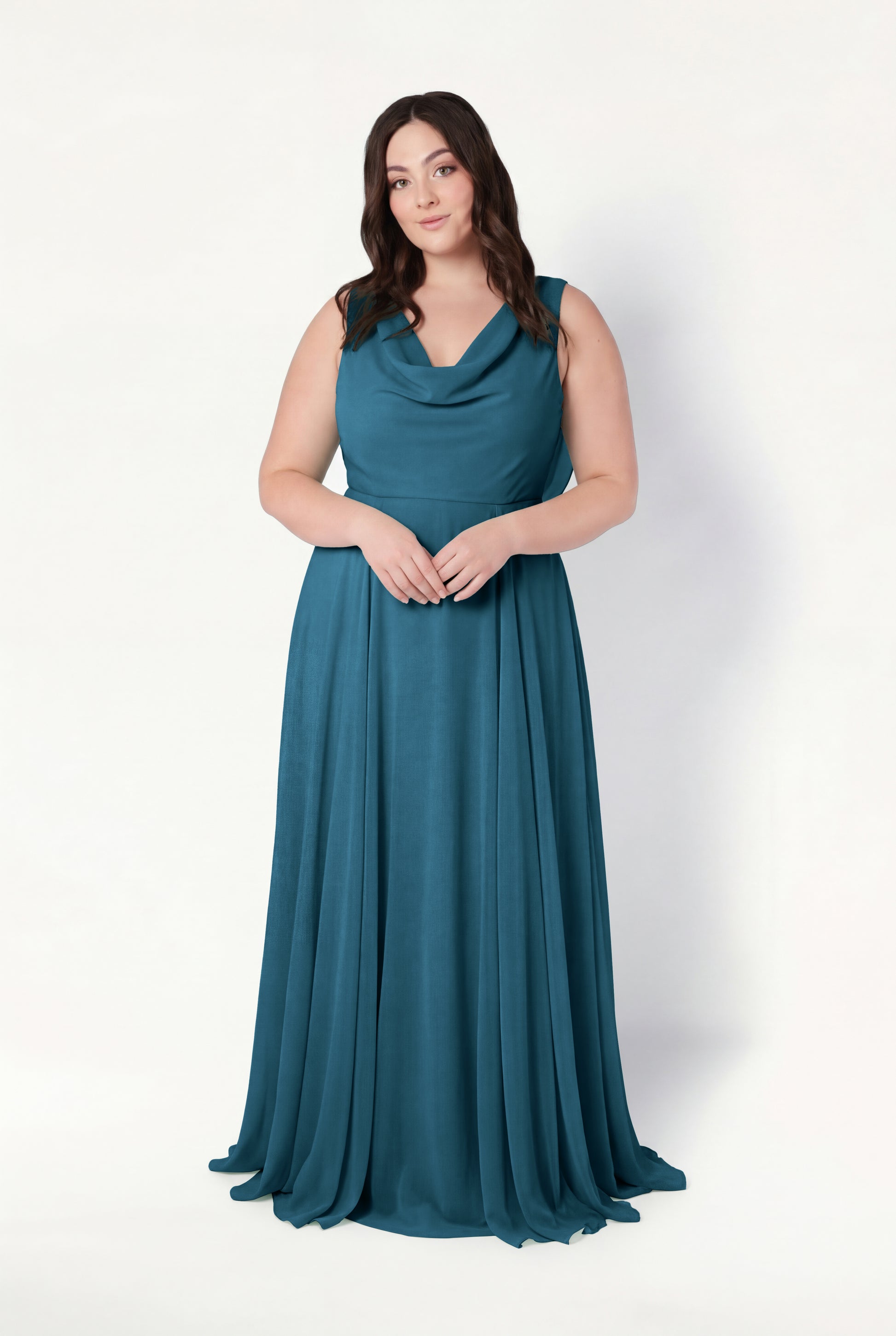 A woman with long brown hair poses against a white background in the TH&TH Athena Bridesmaid Dress in Emerald Dawn, featuring a draped neckline and flowing skirt. Her hands are clasped, expression neutral, exuding timeless elegance.