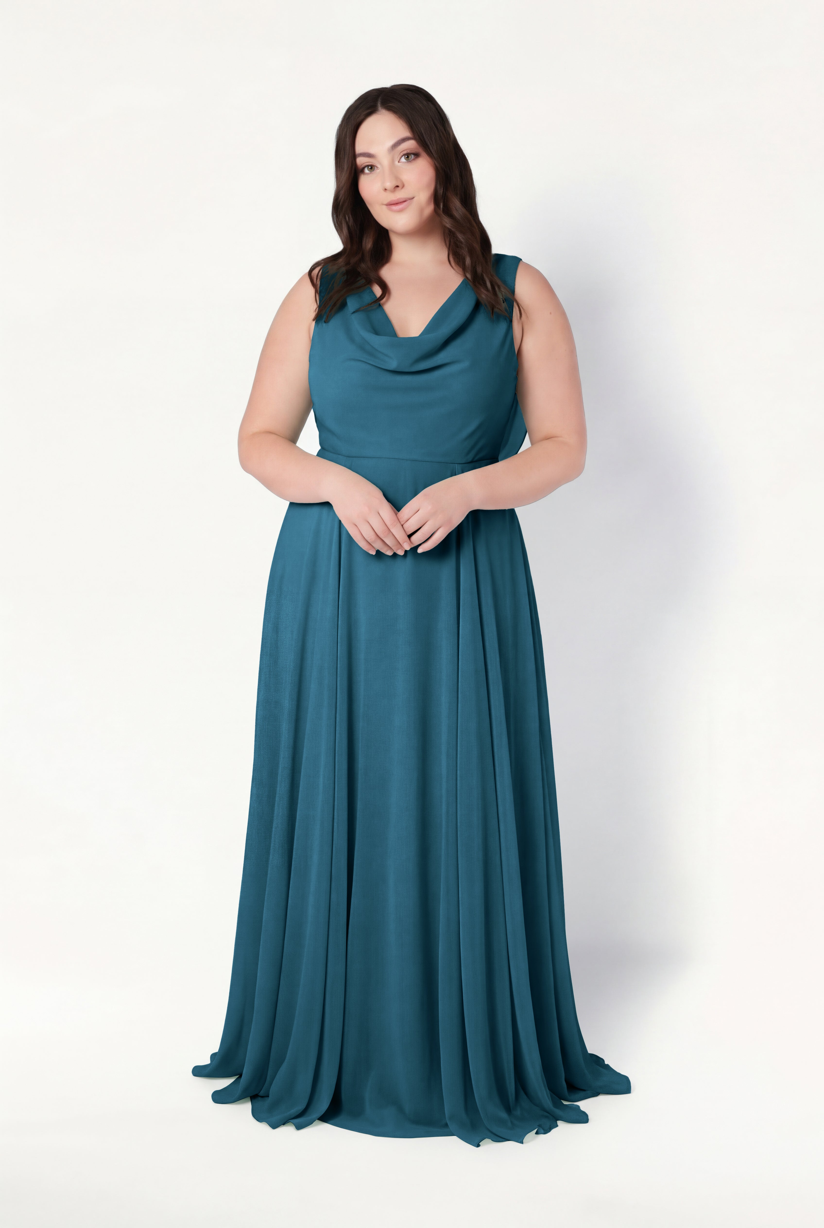 A woman with long brown hair poses against a white background in the TH&TH Athena Bridesmaid Dress in Emerald Dawn, featuring a draped neckline and flowing skirt. Her hands are clasped, expression neutral, exuding timeless elegance.