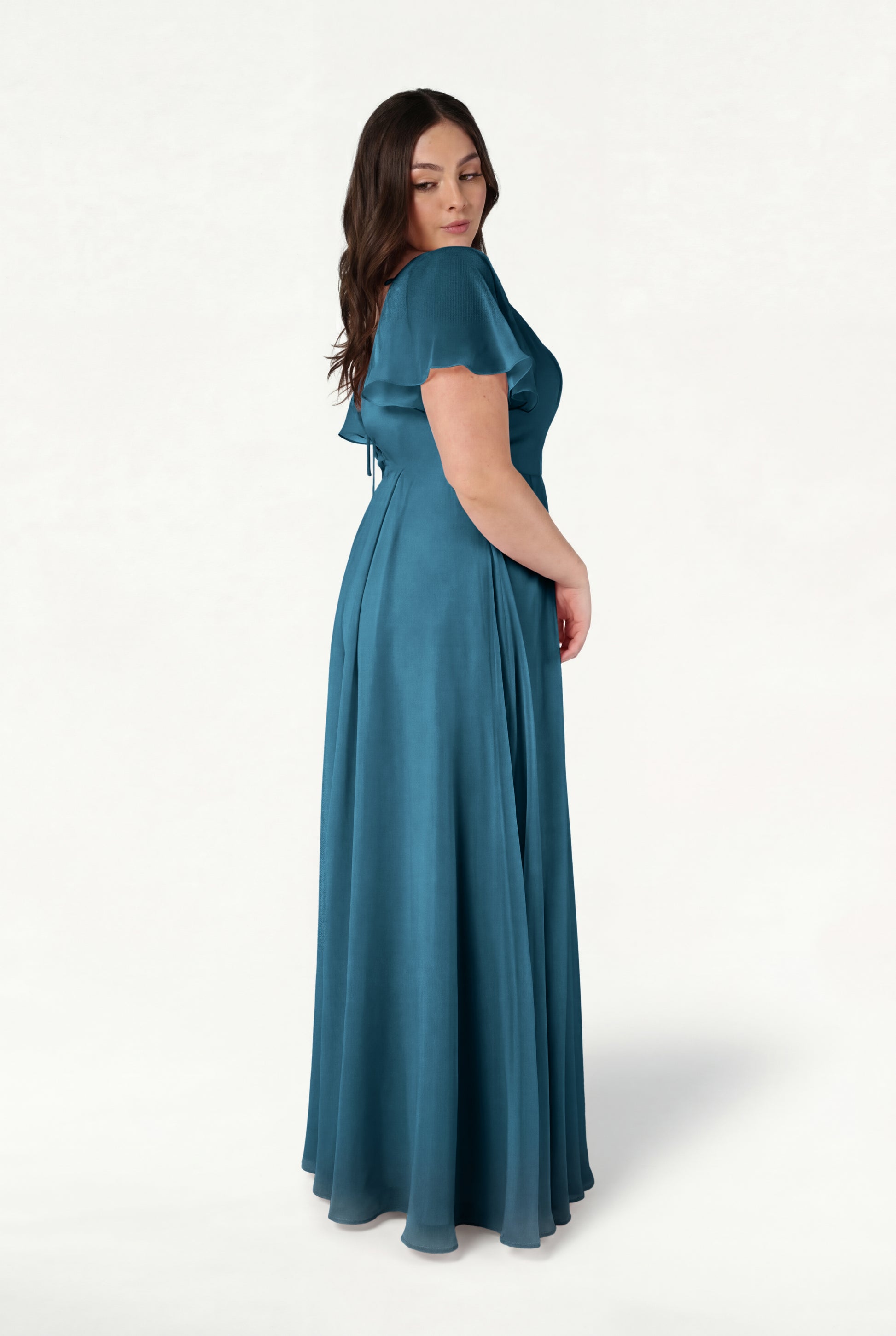 A woman with long brown hair stands sideways against a plain white background, wearing the TH&TH Phoebe Bridesmaid Dress in Emerald Dawn Chiffon—a floor-length, bump-friendly gown—while looking over her shoulder with a relaxed expression.