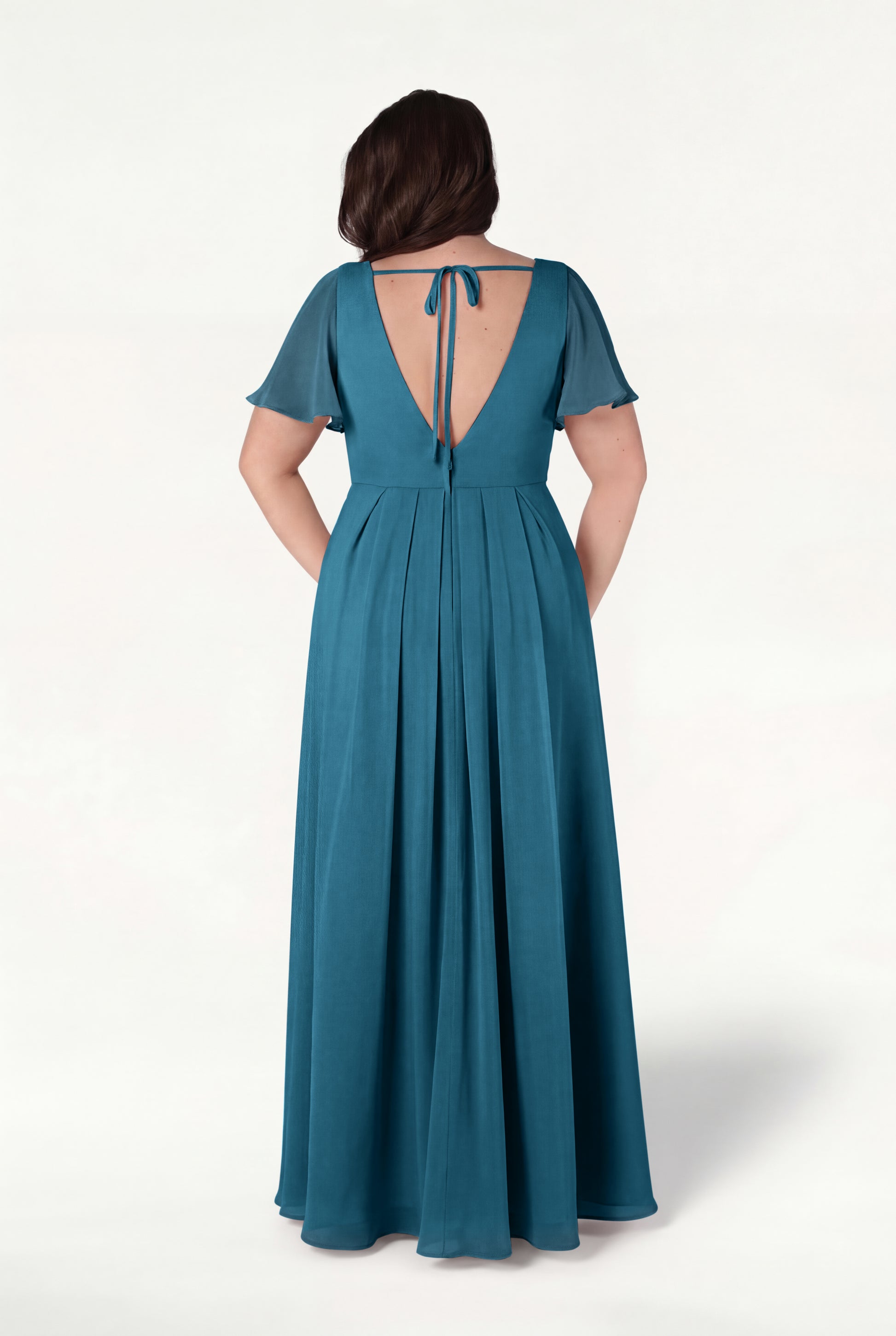A woman faces away in the TH&TH Phoebe Bridesmaid Dress in Emerald Dawn chiffon—a floor-length, bump-friendly gown with short flutter sleeves, pleated skirt, and open tie-back detail—set against a plain white background.