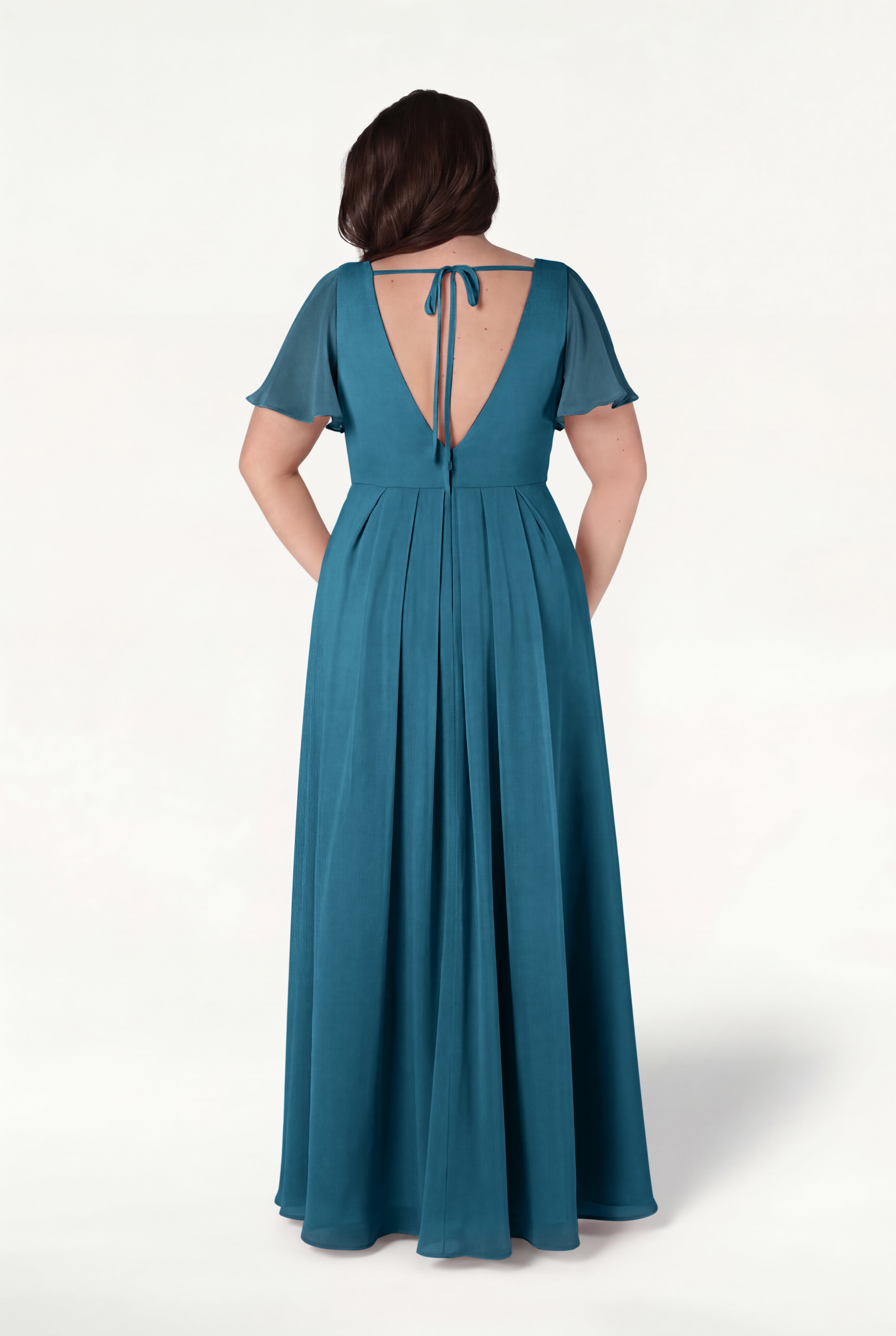 A woman faces away in the TH&TH Phoebe Bridesmaid Dress in Emerald Dawn chiffon—a floor-length, bump-friendly gown with short flutter sleeves, pleated skirt, and open tie-back detail—set against a plain white background.