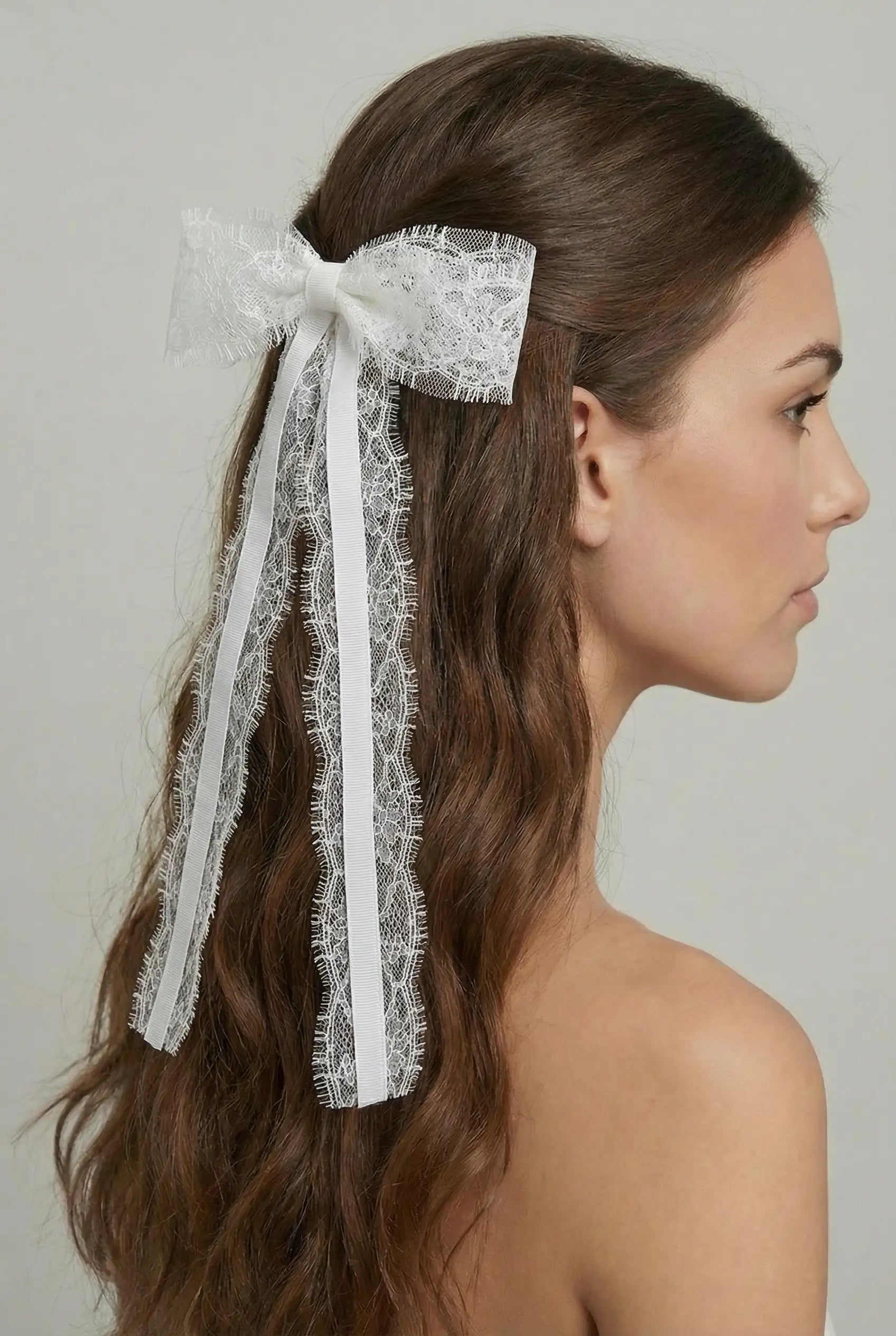 A woman with long, wavy brown hair wears the TH&TH Emmeline Lace Hair Bow—an elegant white Chantilly lace bridal accessory—styled at the back of her head against a plain light gray background.