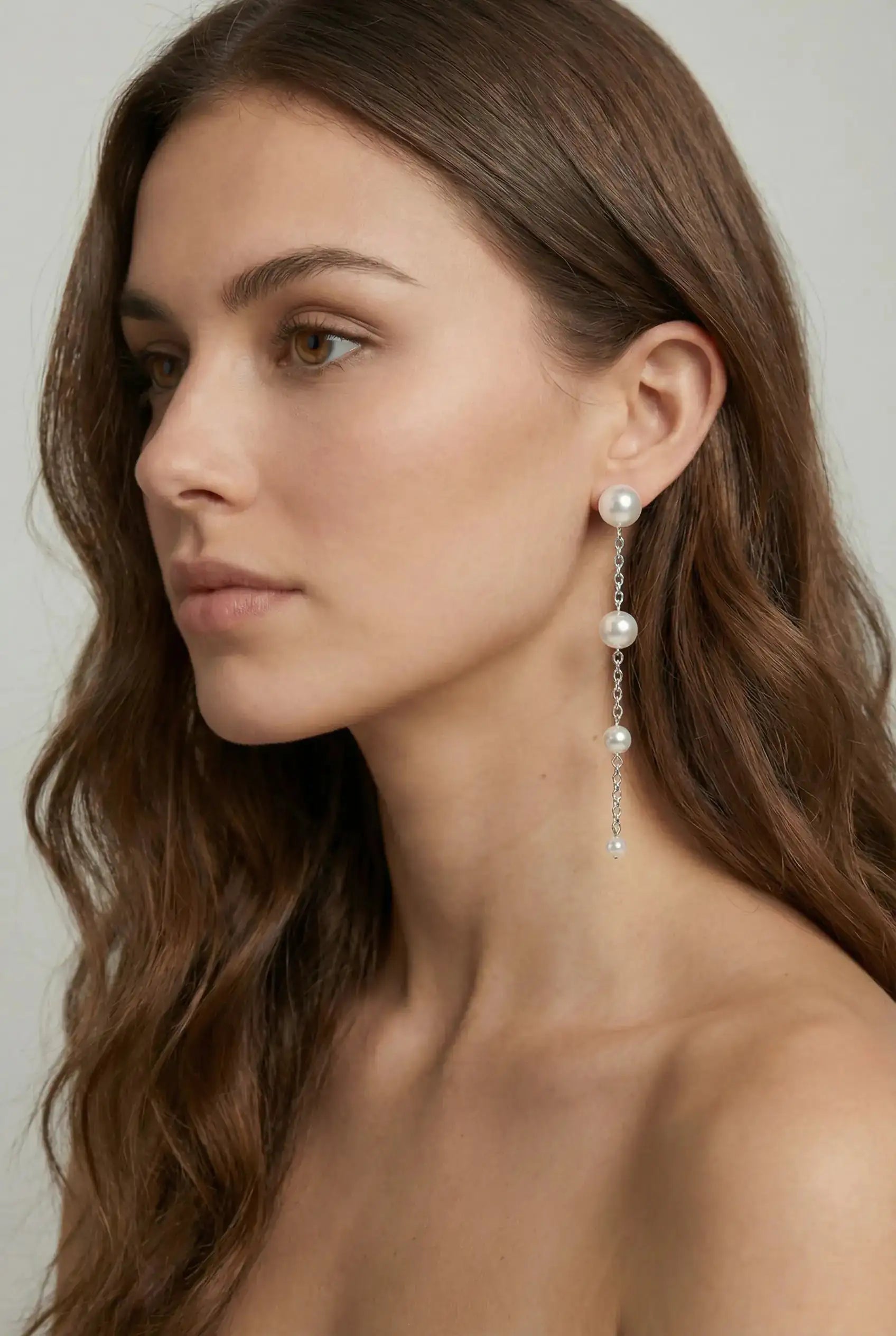 A woman with long brown hair wears TH&TH Empress Pearl Drop Earrings—elegant silver drops featuring three pearls and a small gem. She poses with bare shoulders and a neutral expression against a plain background.