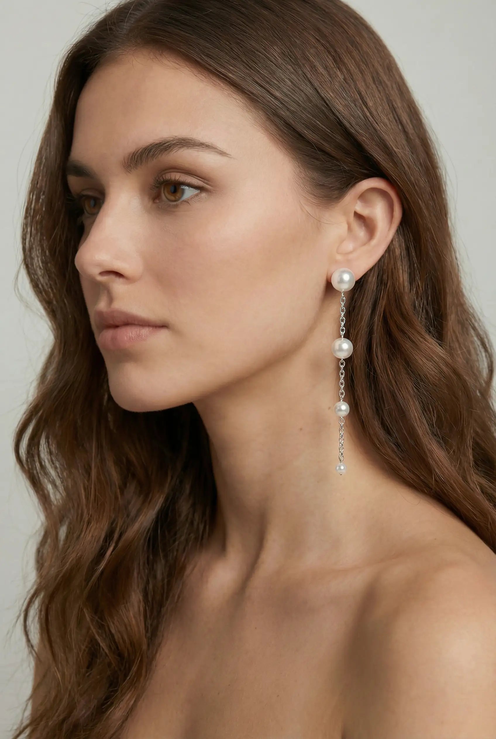 A woman with long brown hair wears TH&TH Empress Pearl Drop Earrings—elegant silver drops featuring three pearls and a small gem. She poses with bare shoulders and a neutral expression against a plain background.
