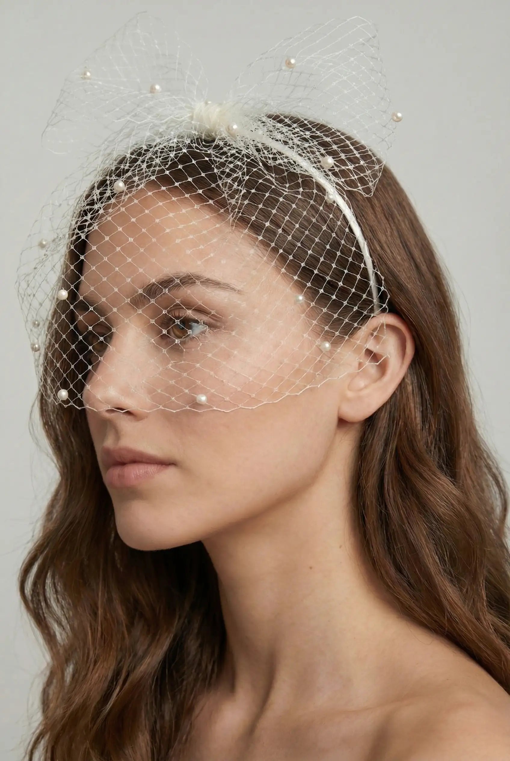 A woman with long brown hair wears the TH&TH Fitzroy Headband with Birdcage, Bow and Scattered Pearls, featuring a large mesh bow and pearls. She faces left against a neutral background, the veil draping over her face.