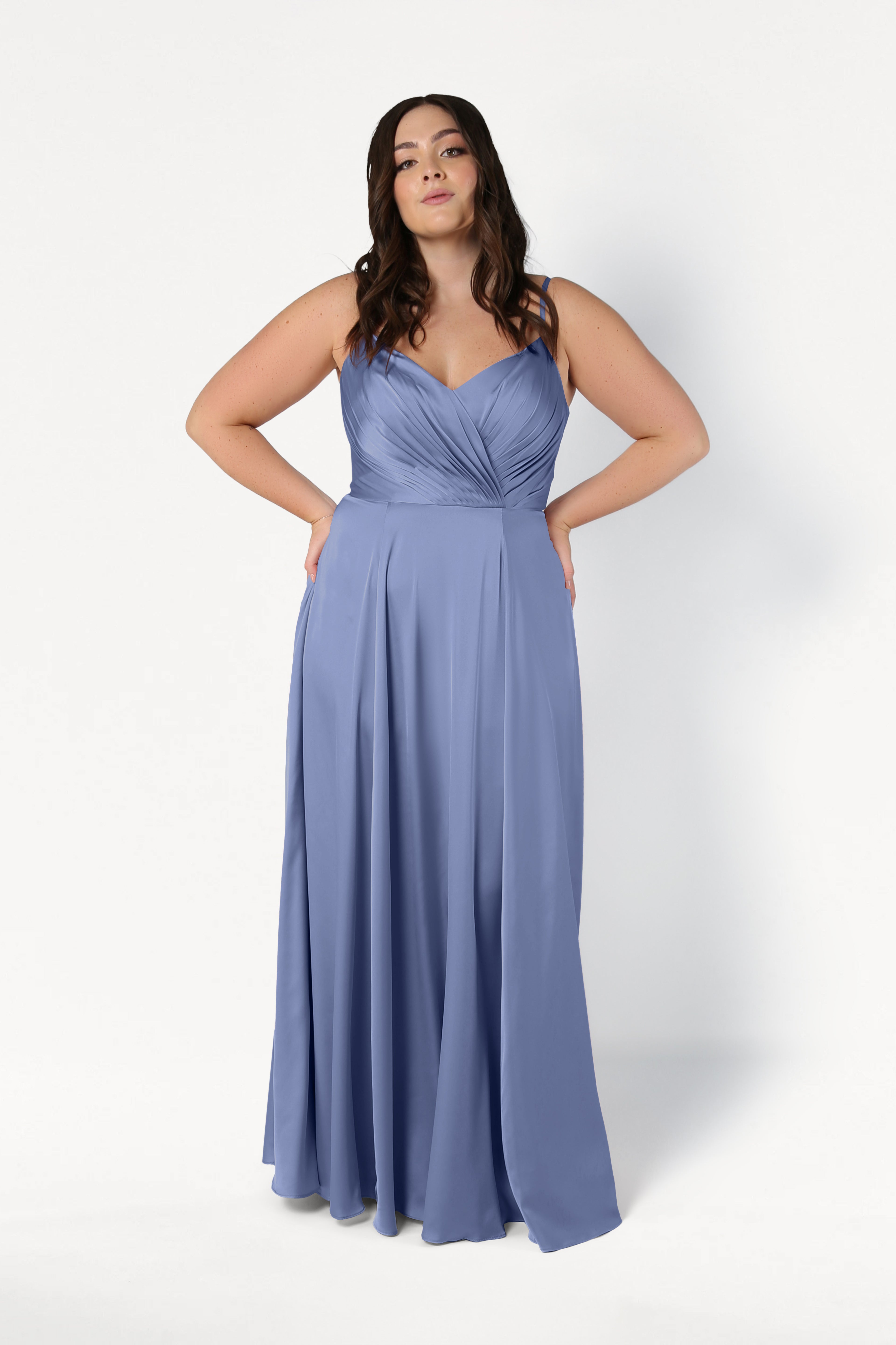 A woman with long, wavy dark hair stands against a white background wearing the TH&TH Isla Bridesmaid Dress in French Blue Satin, featuring spaghetti straps and a pleated bodice. Her hands rest on her hips.