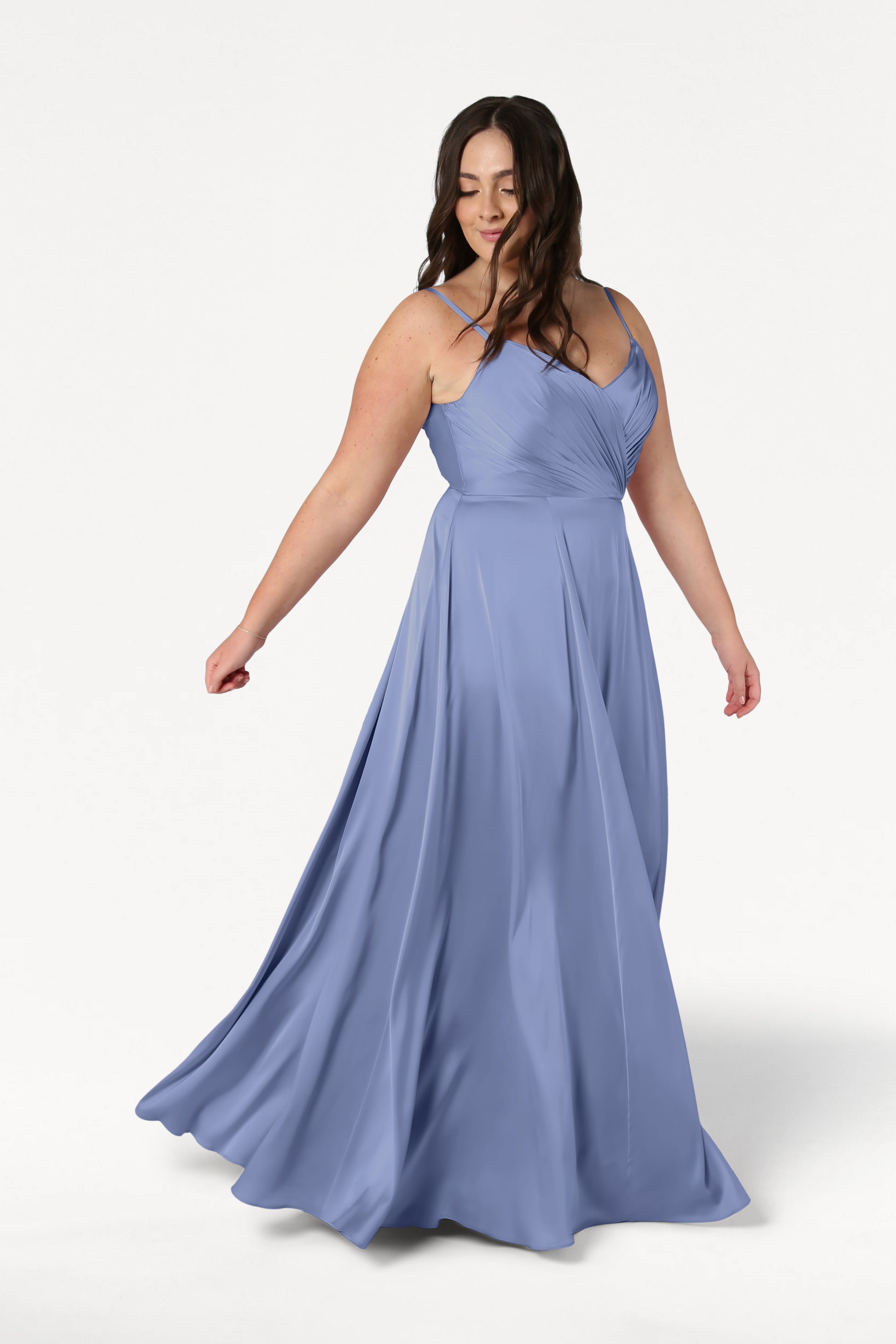 A woman with long brown hair twirls gracefully in the TH&TH Isla Bridesmaid Dress in French Blue Satin, featuring a flowing silhouette and pleated bodice, set against a plain white background.