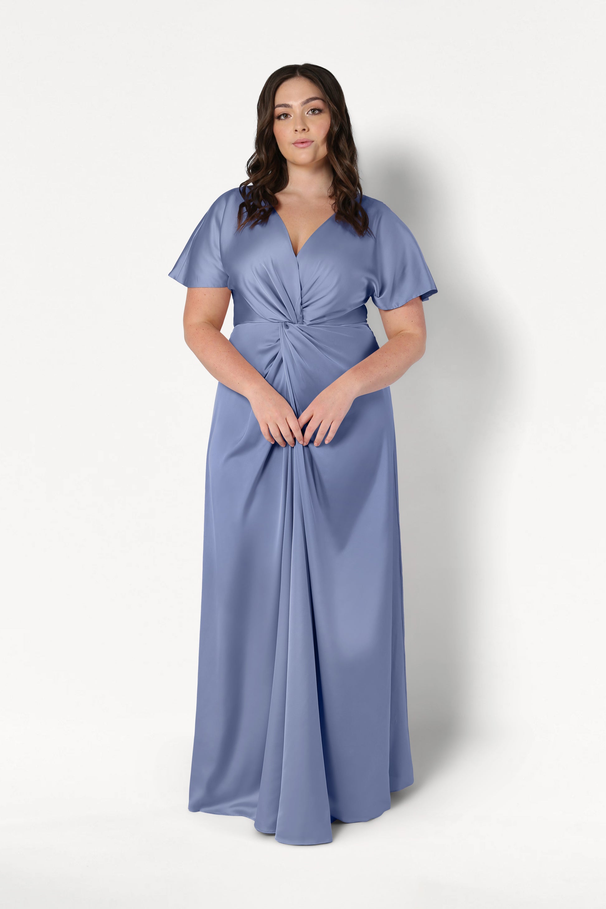 A woman with long dark hair wears the TH&TH Camilla Bridesmaid Dress in French Blue Satin—a floor-length, short-sleeved gown with a twist-knot bodice—standing facing forward against a plain white background.