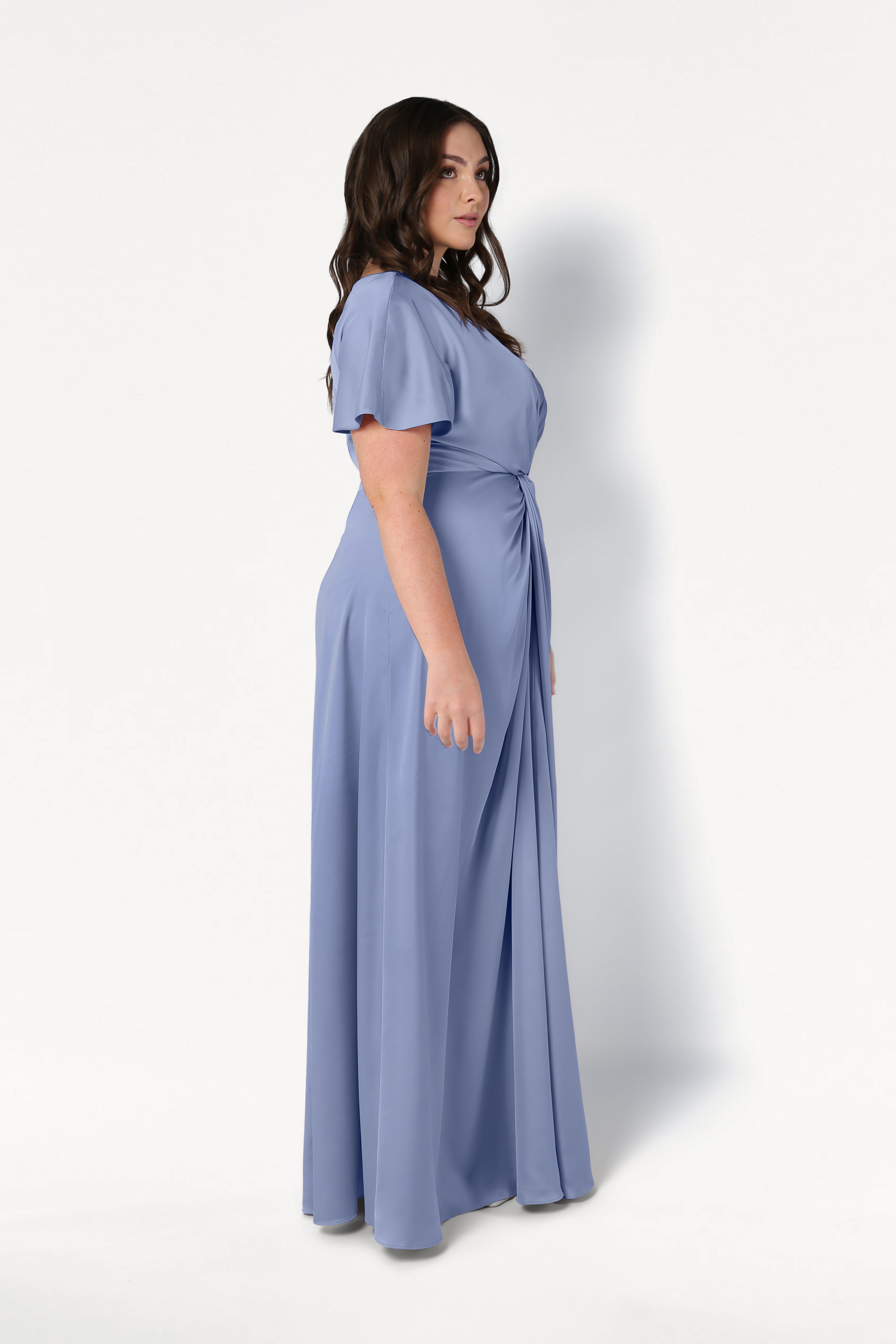 A woman with long brown hair stands sideways in the TH&TH Camilla Bridesmaid Dress, a size-inclusive, floor-length French Blue satin gown with short sleeves and gathered waist detail, set against a plain white background.