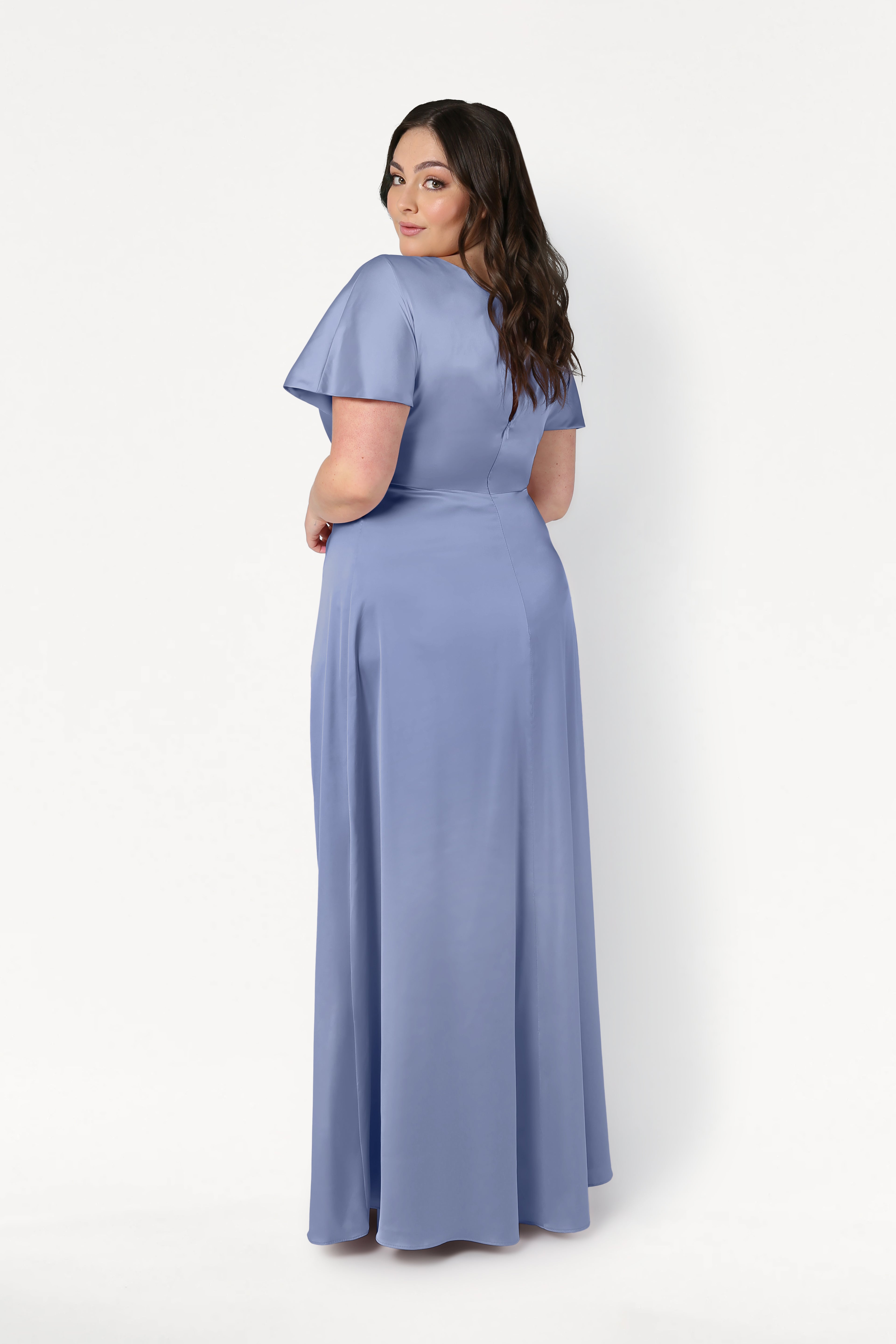 A woman with long dark hair stands facing away, looking over her shoulder in the TH&TH Camilla Bridesmaid Dress in French Blue Satin—a floor-length, short-sleeve gown with a twist-knot bodice—set against a plain white background.