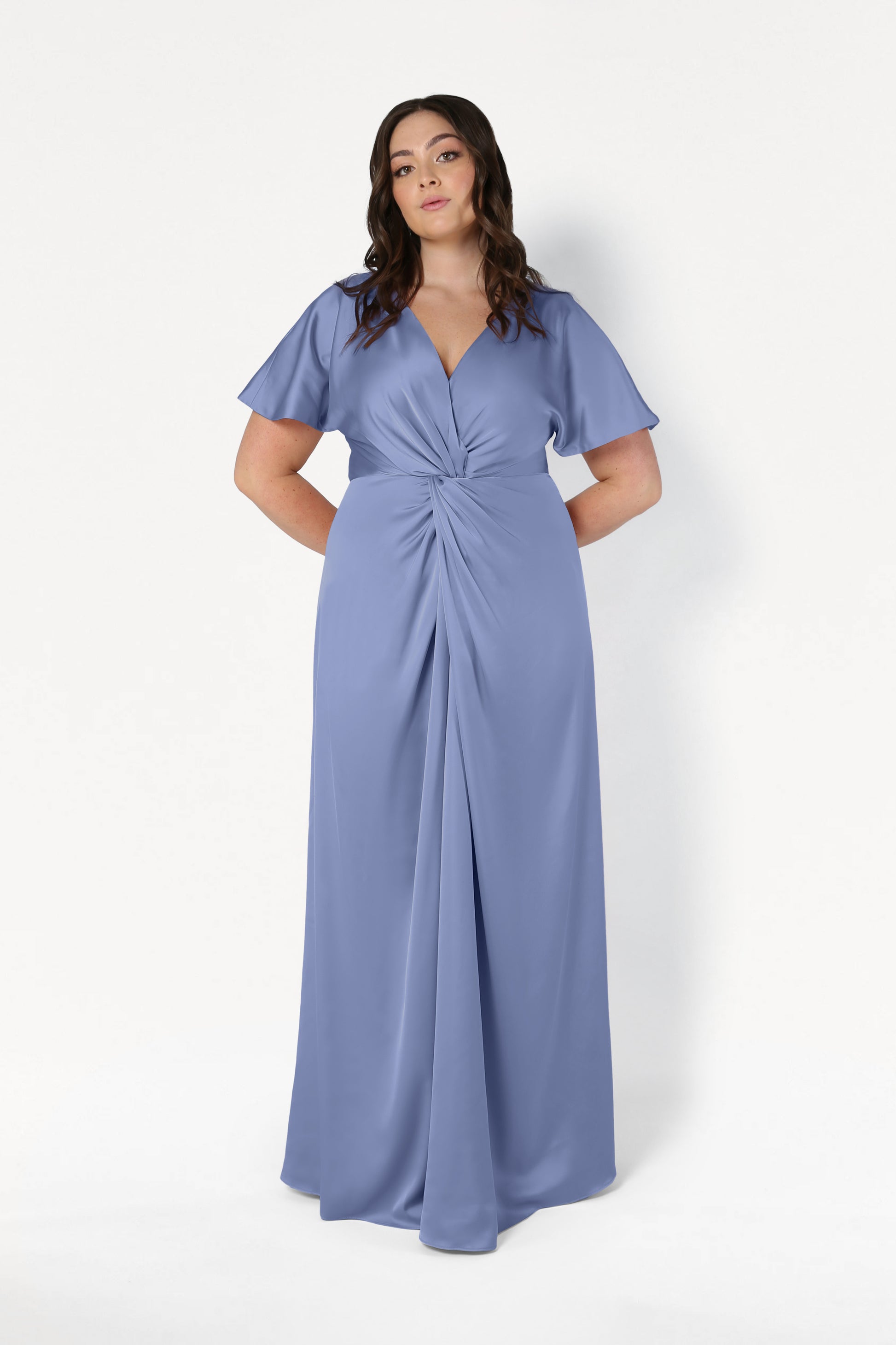 A woman with wavy dark hair and a neutral expression stands against a plain white background wearing the TH&TH Camilla Bridesmaid Dress in French Blue Satin—a size-inclusive, floor-length gown featuring a V-neck and twist-knot bodice.