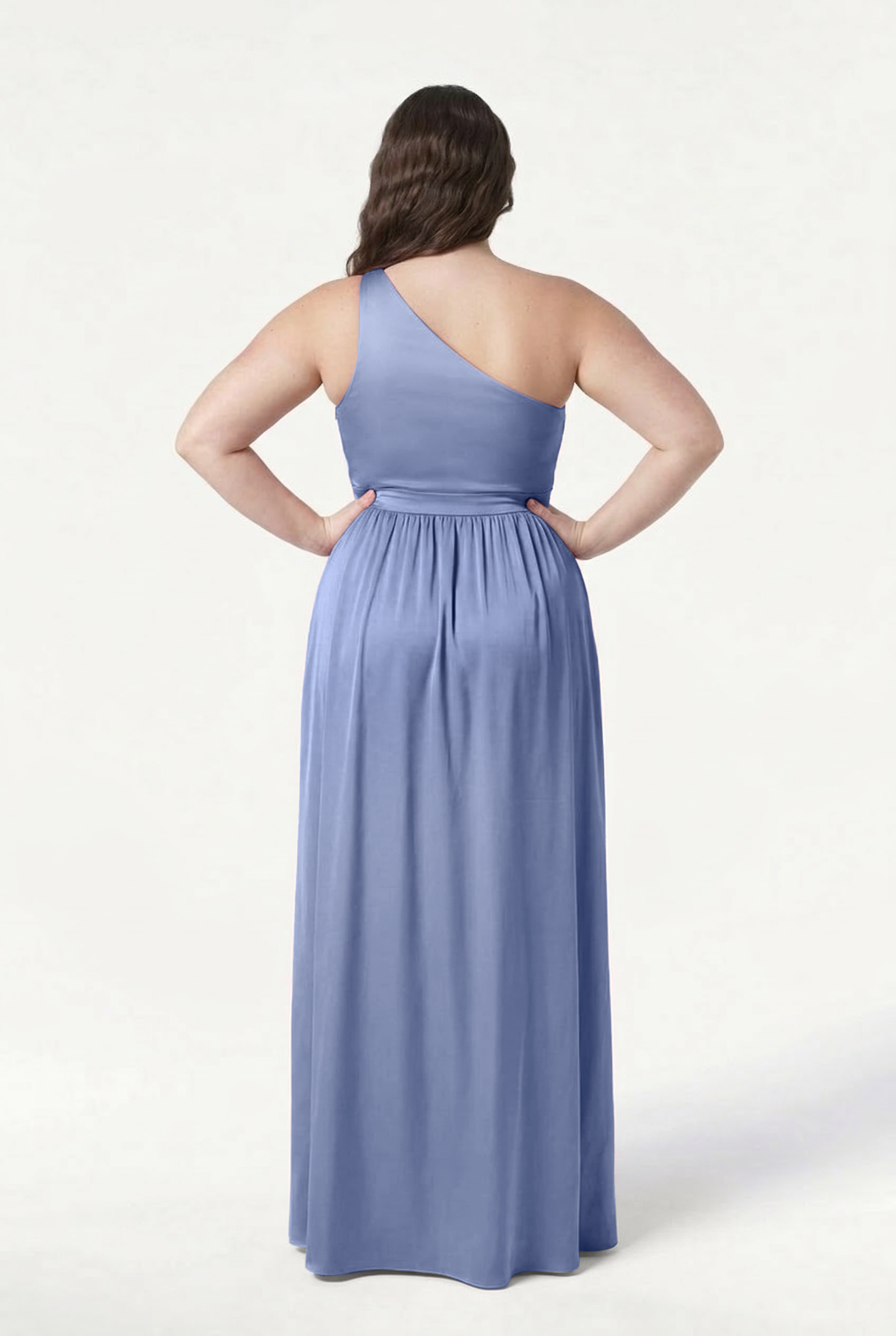 A woman stands facing away, hands on hips, wearing the TH&TH Greta Bridesmaid Dress in French Blue Satin. This elegant gown features a gathered waist and one-shoulder design, set against a plain white background.