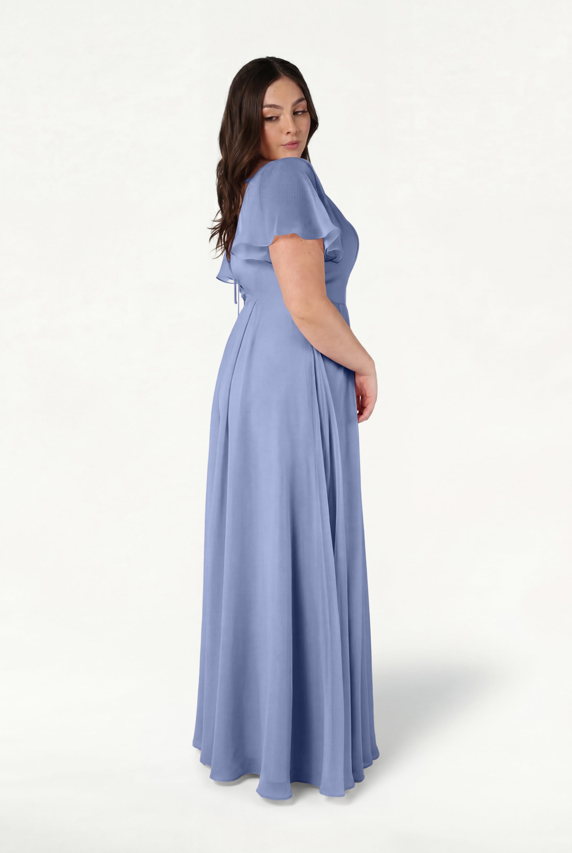 A woman with long brown hair stands sideways against a plain white background, wearing the TH&TH Phoebe Bridesmaid Dress in French Blue Chiffon—a flowing, short-sleeve gown.