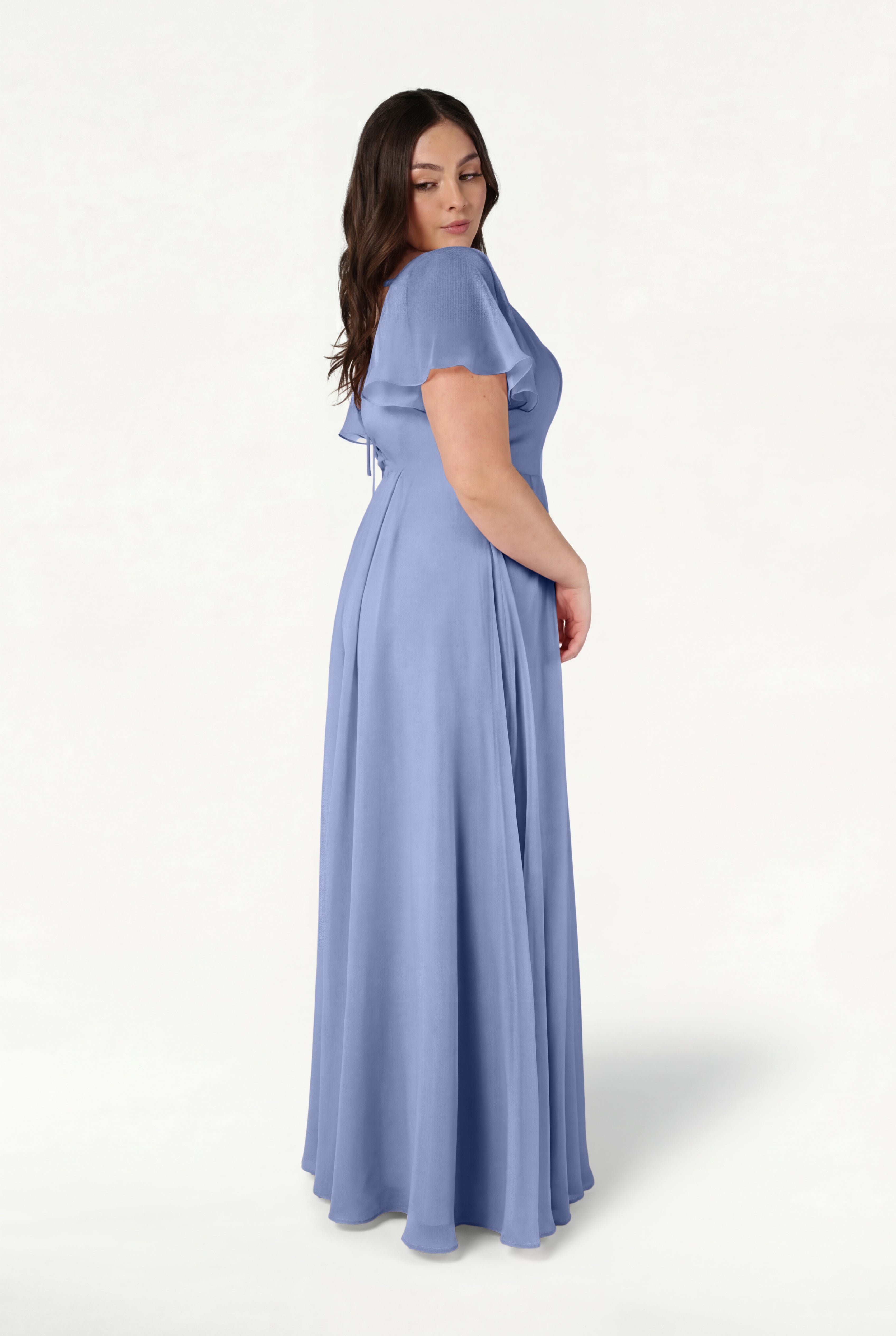A woman with long brown hair stands sideways against a plain white background, wearing the TH&TH Phoebe Bridesmaid Dress in French Blue Chiffon—a flowing, short-sleeve gown.