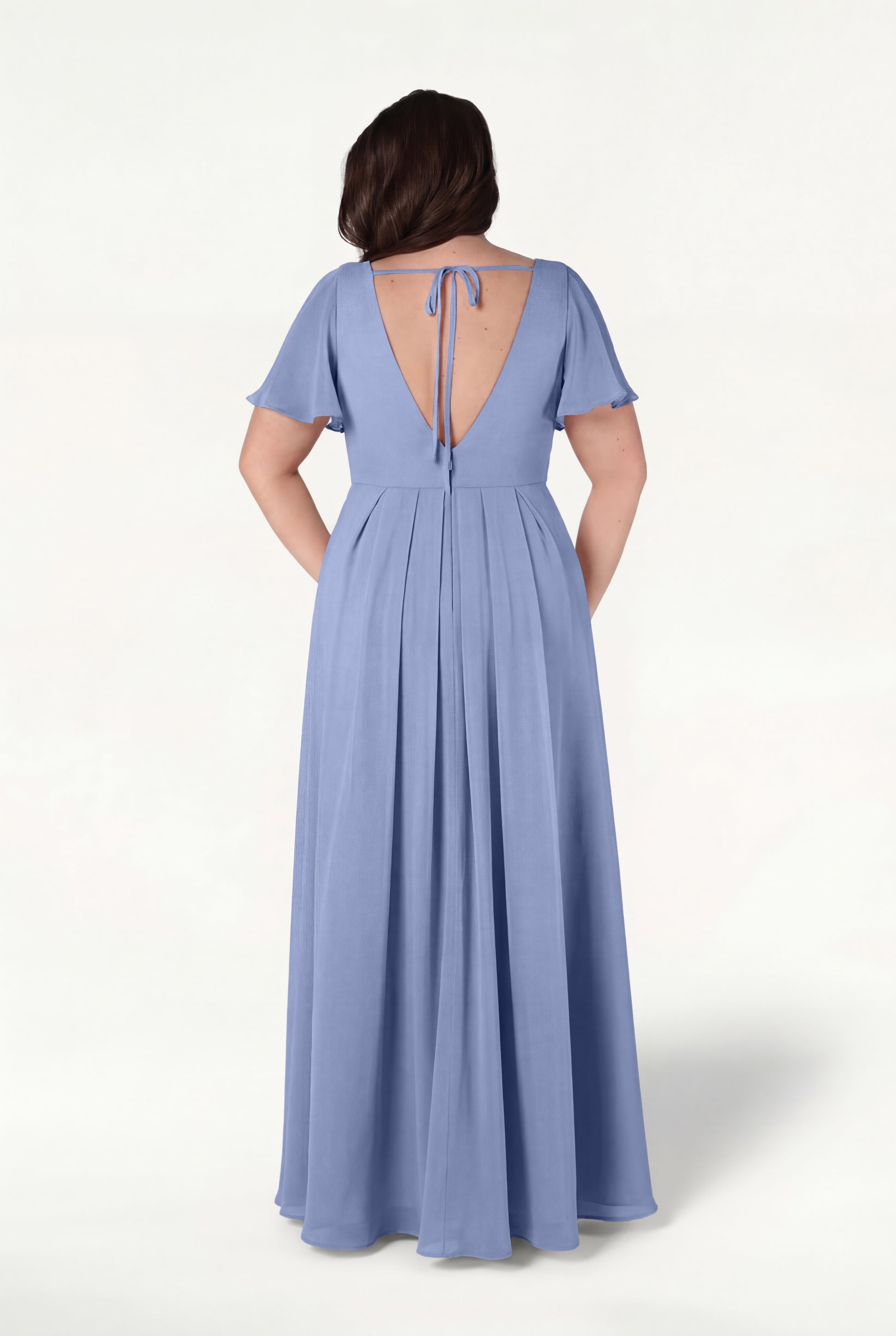 A woman stands against a plain light background, hands in her pockets, wearing the TH&TH Phoebe Bridesmaid Dress in French Blue Chiffon—a floor-length gown with short sleeves and a deep V-back tied at the top with a string.