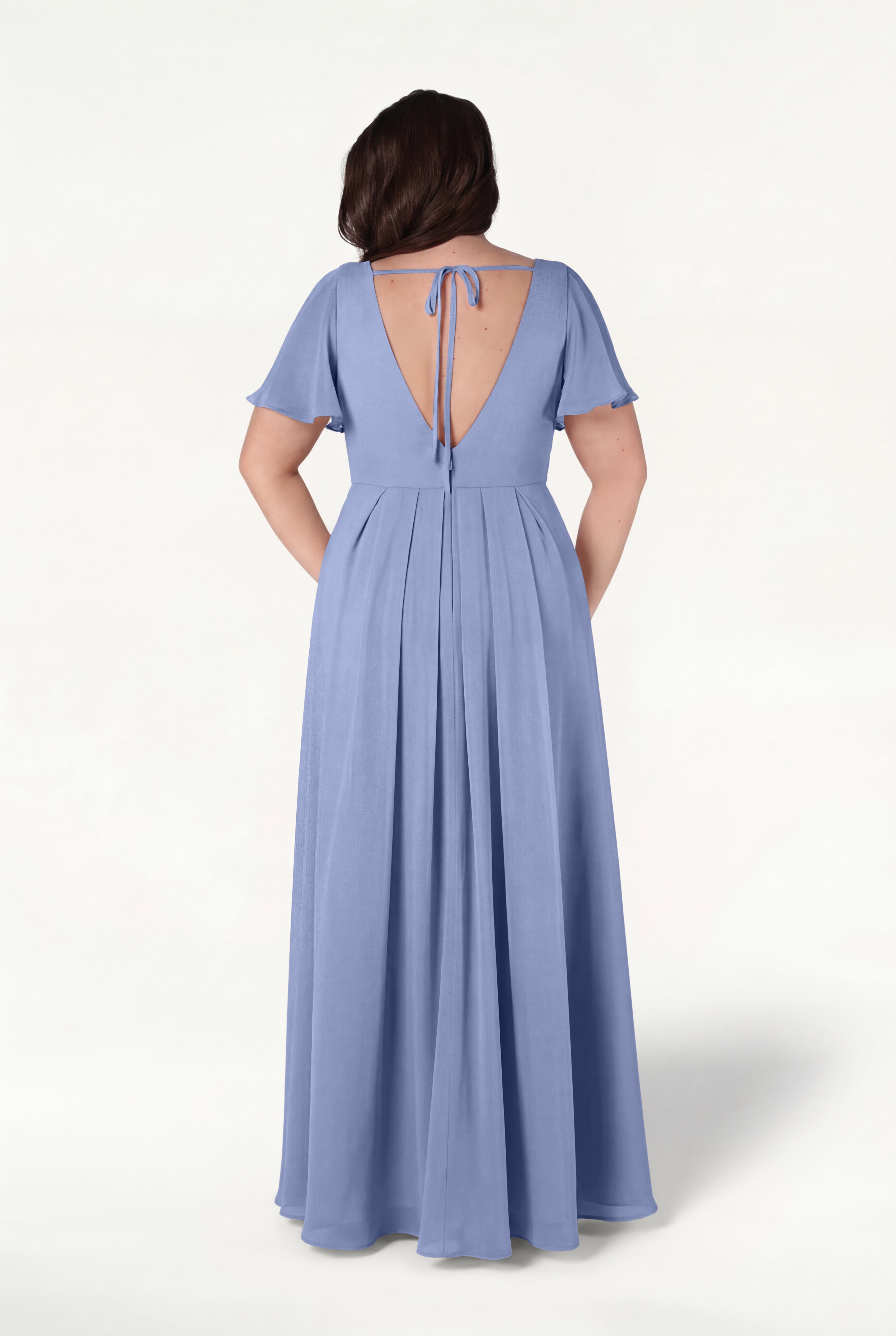 A woman stands against a plain light background, hands in her pockets, wearing the TH&TH Phoebe Bridesmaid Dress in French Blue Chiffon—a floor-length gown with short sleeves and a deep V-back tied at the top with a string.