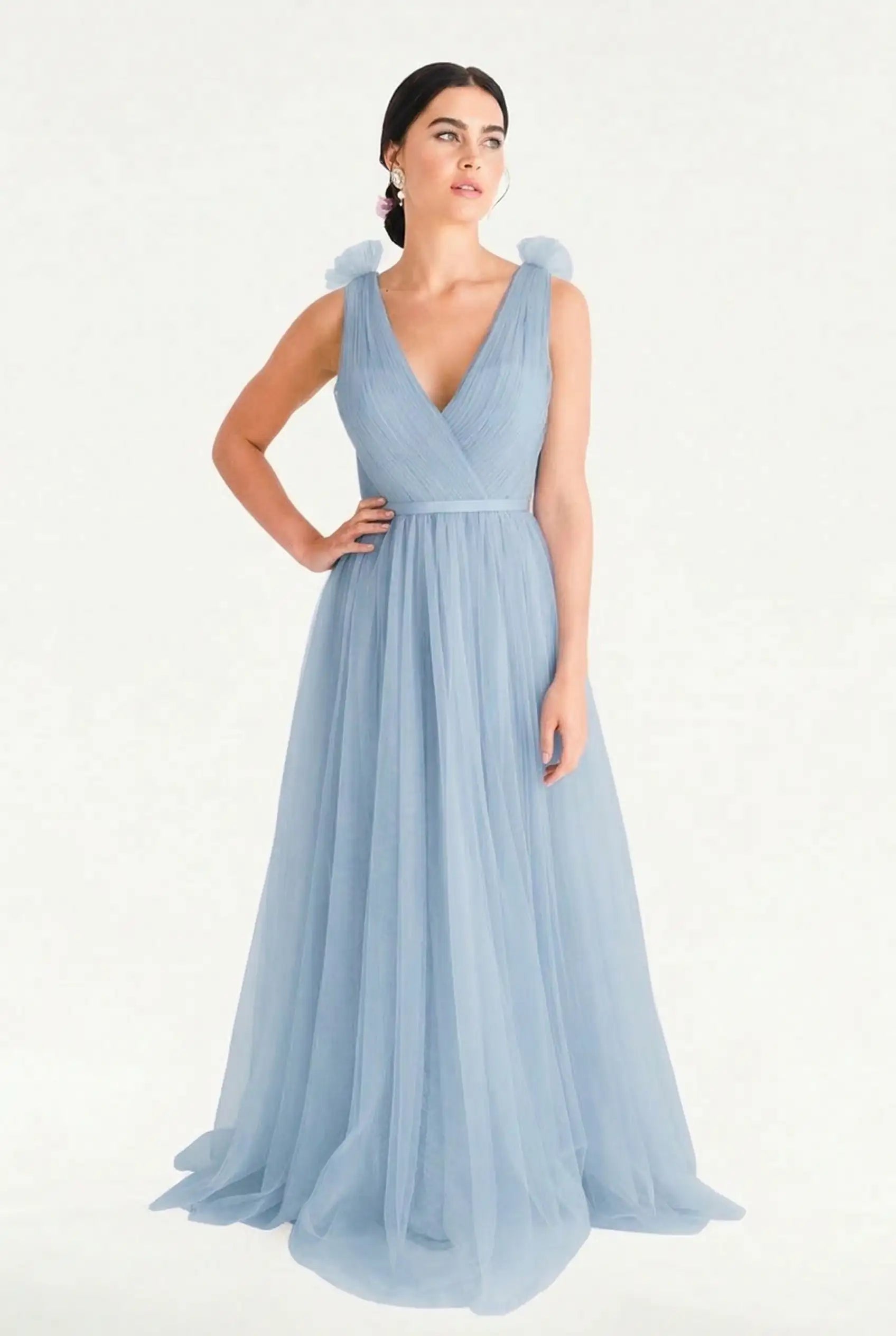 A woman models the TH&TH Grace Bridesmaid Dress in Dusty Blue Tulle, a sleeveless, floor-length gown with a V-neckline and soft shoulder details, standing against a white background with one hand on her hip.