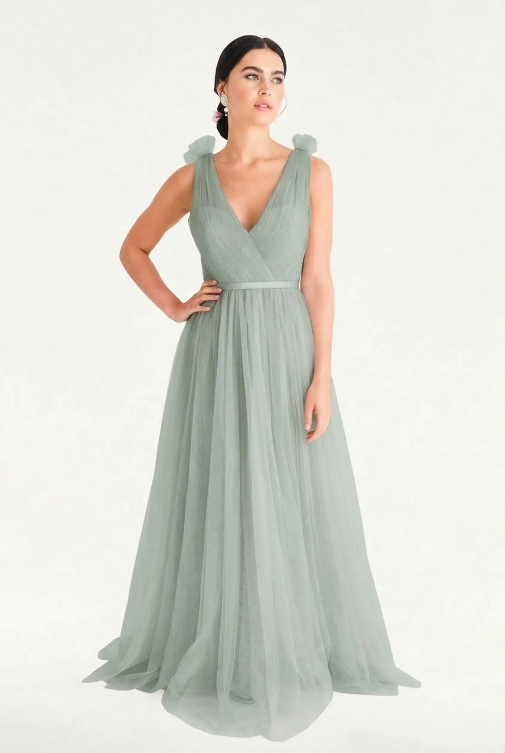 A woman models the TH&TH Grace Bridesmaid Dress in Dusty Eucalyptus Tulle, featuring a long, flowing silhouette with a deep V-neckline and gathered tulle shoulders, against a plain white background.