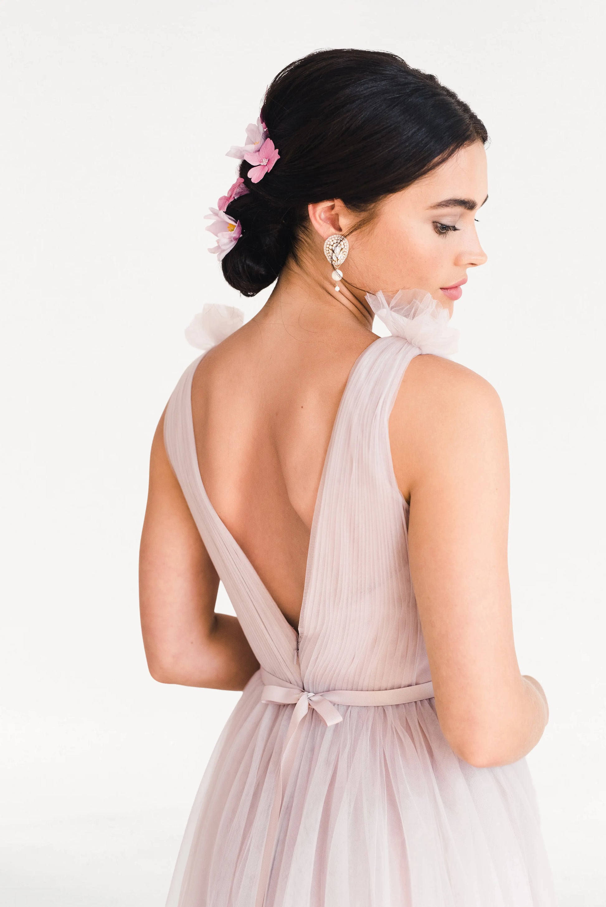 A woman wears the TH&TH Grace Bridesmaid Dress in Smoked Blush Tulle, featuring a sleeveless design and deep V-back. Her dark updo with pink flowers enhances the romantic look—ideal for bridal parties against a white background.