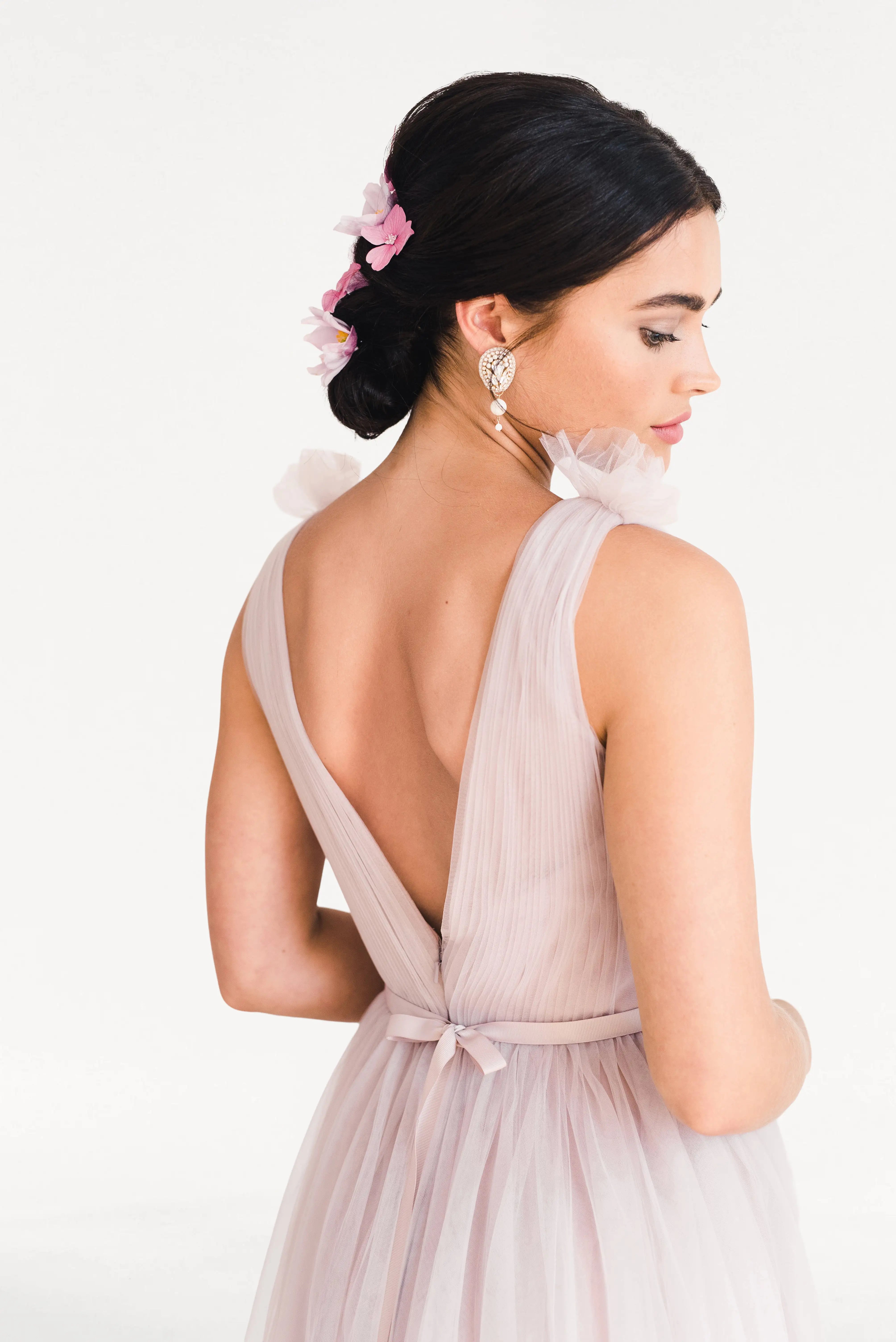 A woman wears the TH&TH Grace Bridesmaid Dress in Smoked Blush Tulle, featuring a sleeveless design and deep V-back. Her dark updo with pink flowers enhances the romantic look—ideal for bridal parties against a white background.