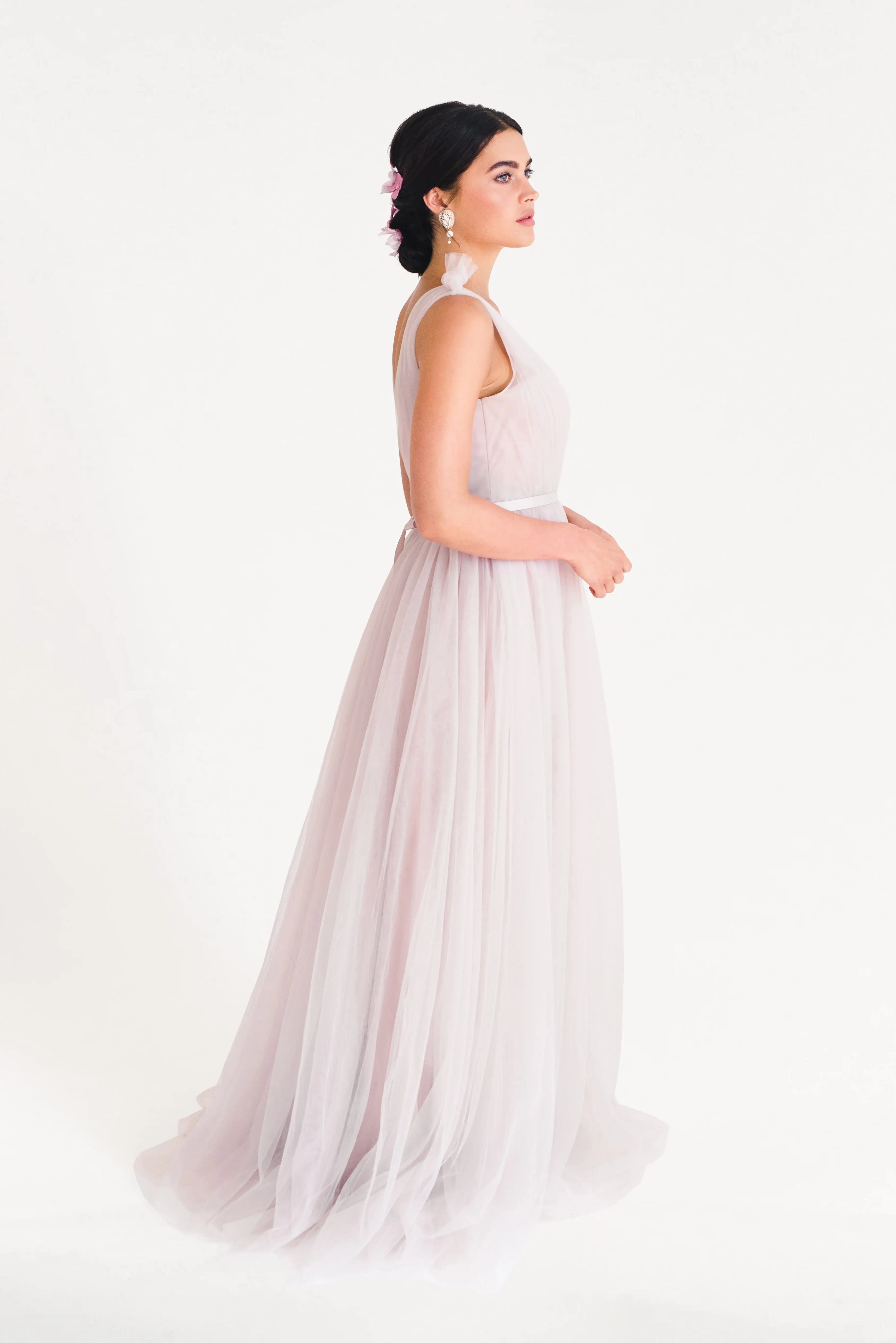 A woman stands in profile against a white backdrop, wearing the TH&TH Grace Bridesmaid Dress in smoked blush tulle—an elegant choice with an updo and floral accents, perfect for a romantic bridal party or as a bridesmaid dress.