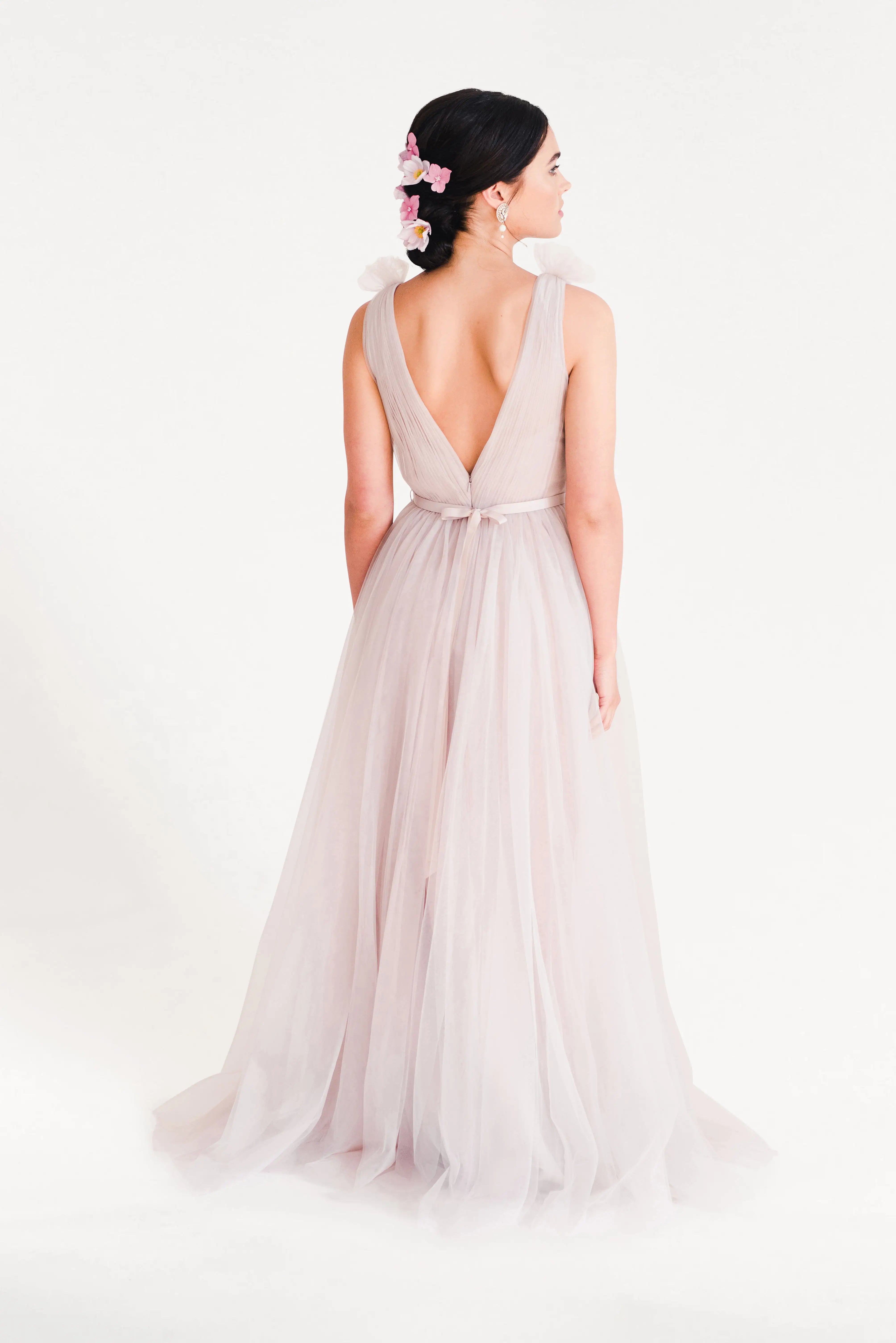 A woman wears the TH&TH Grace Bridesmaid Dress in Smoked Blush Tulle. Her dark hair is styled up with pink flowers, creating a romantic bridal look against a plain white background.