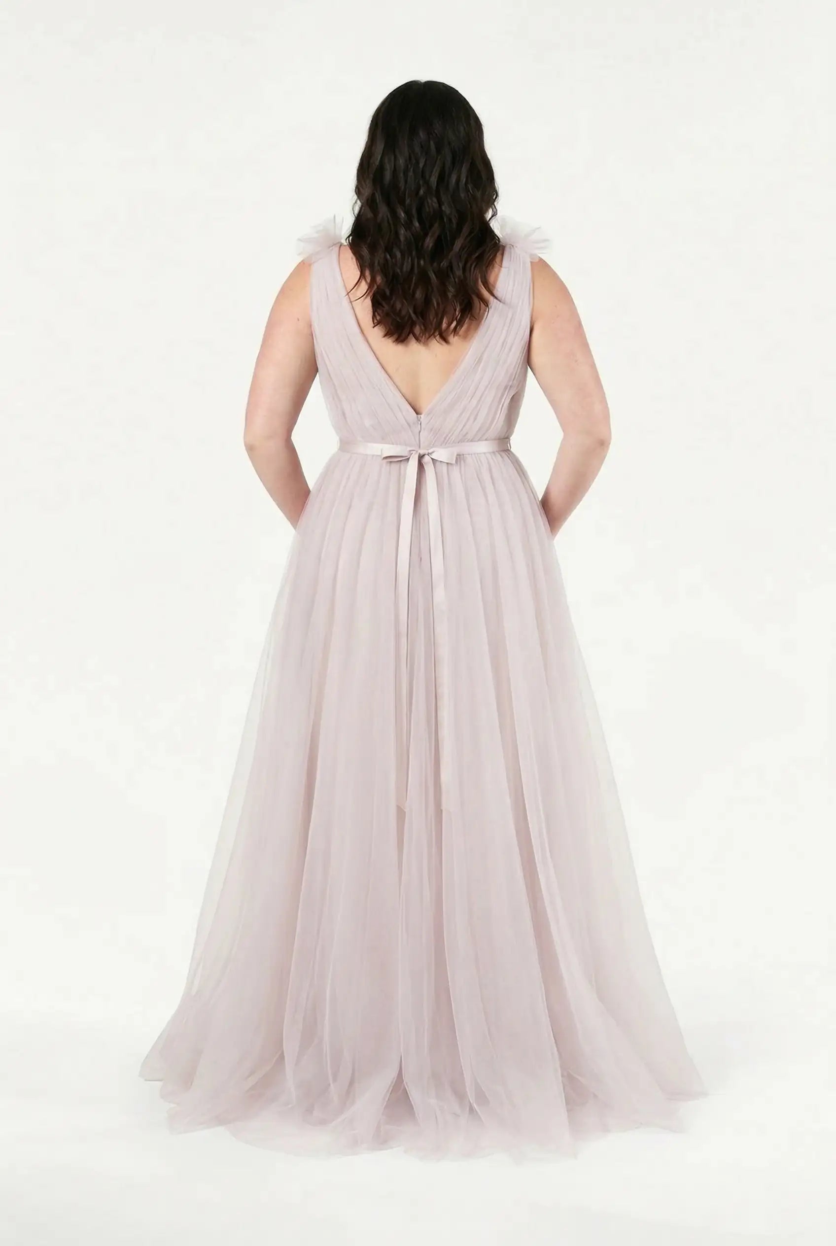 A woman faces away in the TH&TH Grace Bridesmaid Dress in Smoked Blush Tulle, featuring a V-back, ribbon waist tie, and soft ruffle shoulders, set against a white background.