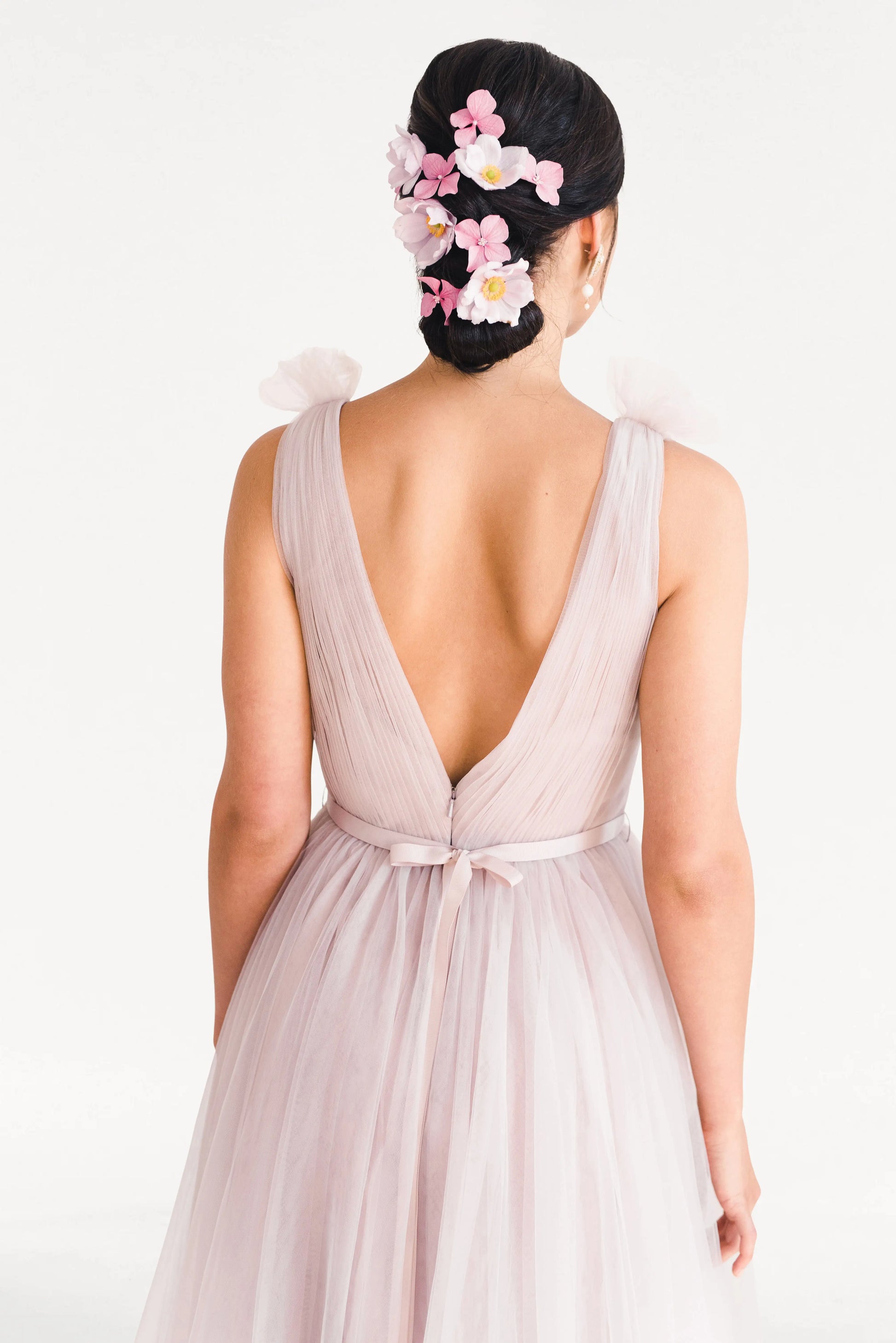 A woman faces away in the TH&TH Grace Bridesmaid Dress in Smoked Blush Tulle, a light pink sleeveless gown with a deep V back. Her dark hair is in a low bun adorned with pink and white flower clips, against a plain white background.